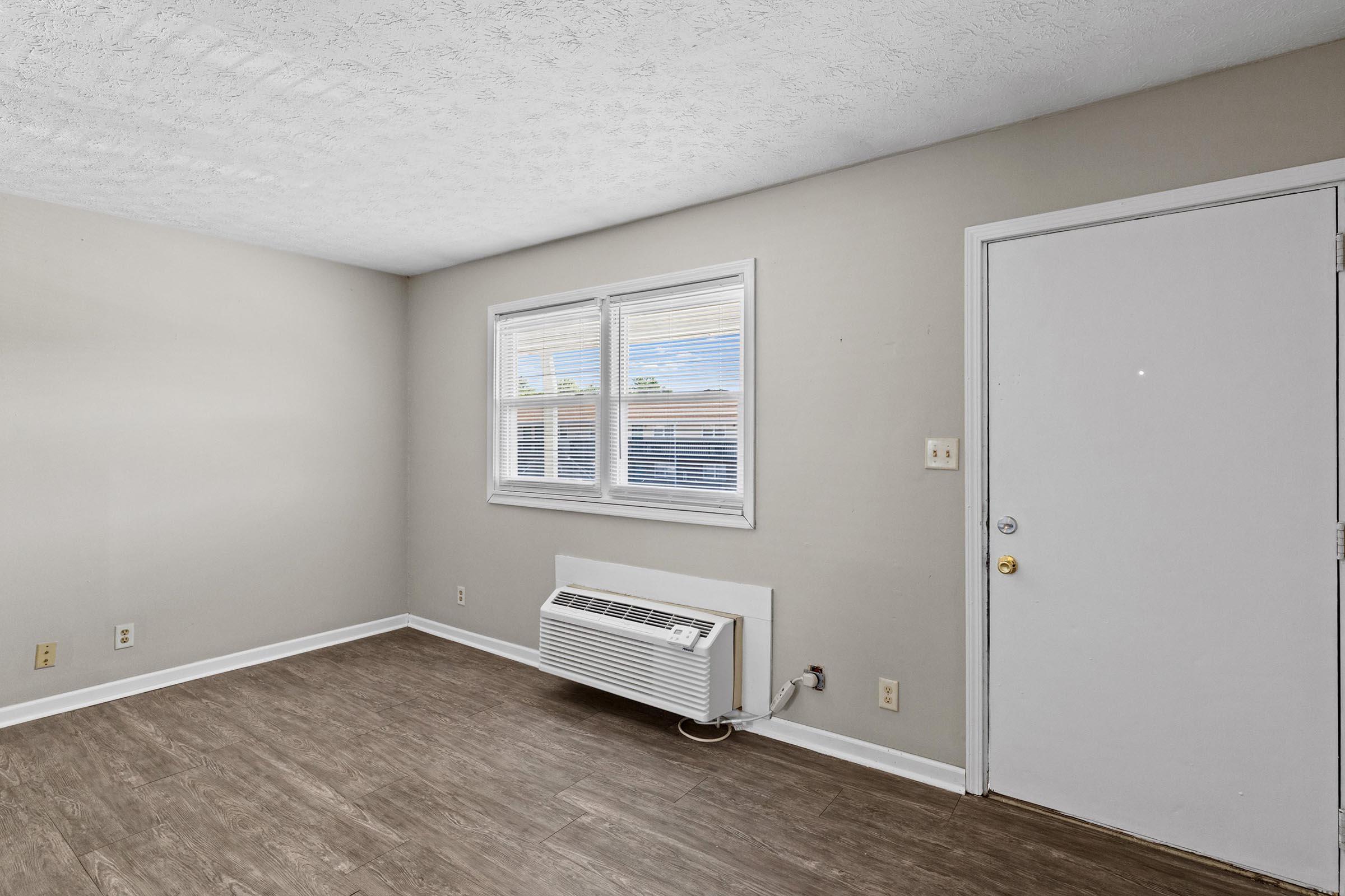 A light-colored room with a textured ceiling and hardwood-style flooring. There are two windows with white blinds on the left side, and a white wall-mounted air conditioning unit beneath them. The entrance door is on the right side, and the walls are painted a neutral color. The overall atmosphere is bright and airy.