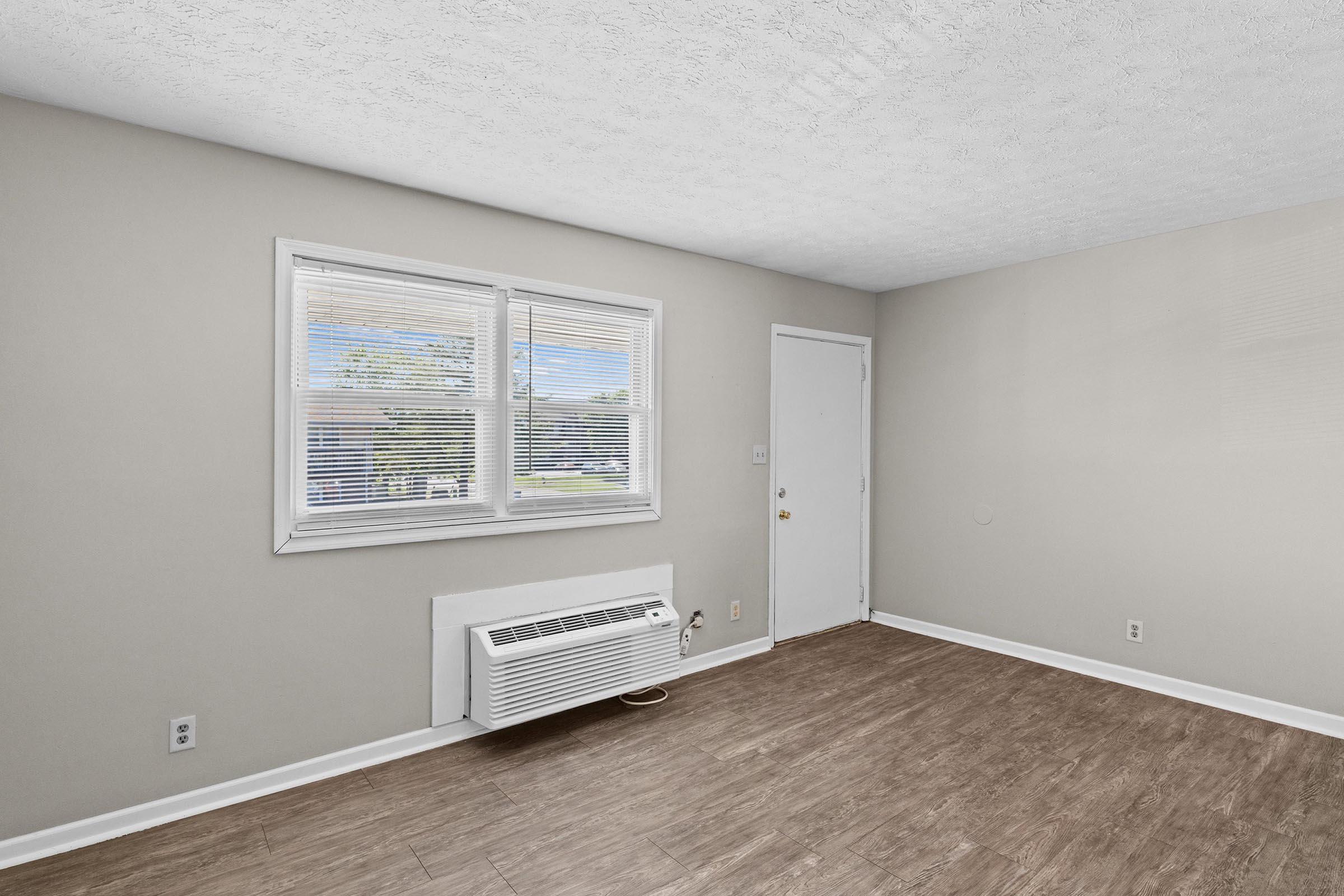 A spacious, light-filled room with a neutral wall color and a wood-like floor. There is a large window allowing natural light to enter, and an air conditioning unit mounted below it. A closed door is visible on the right side, providing access to another area. The overall atmosphere is clean and minimalistic.