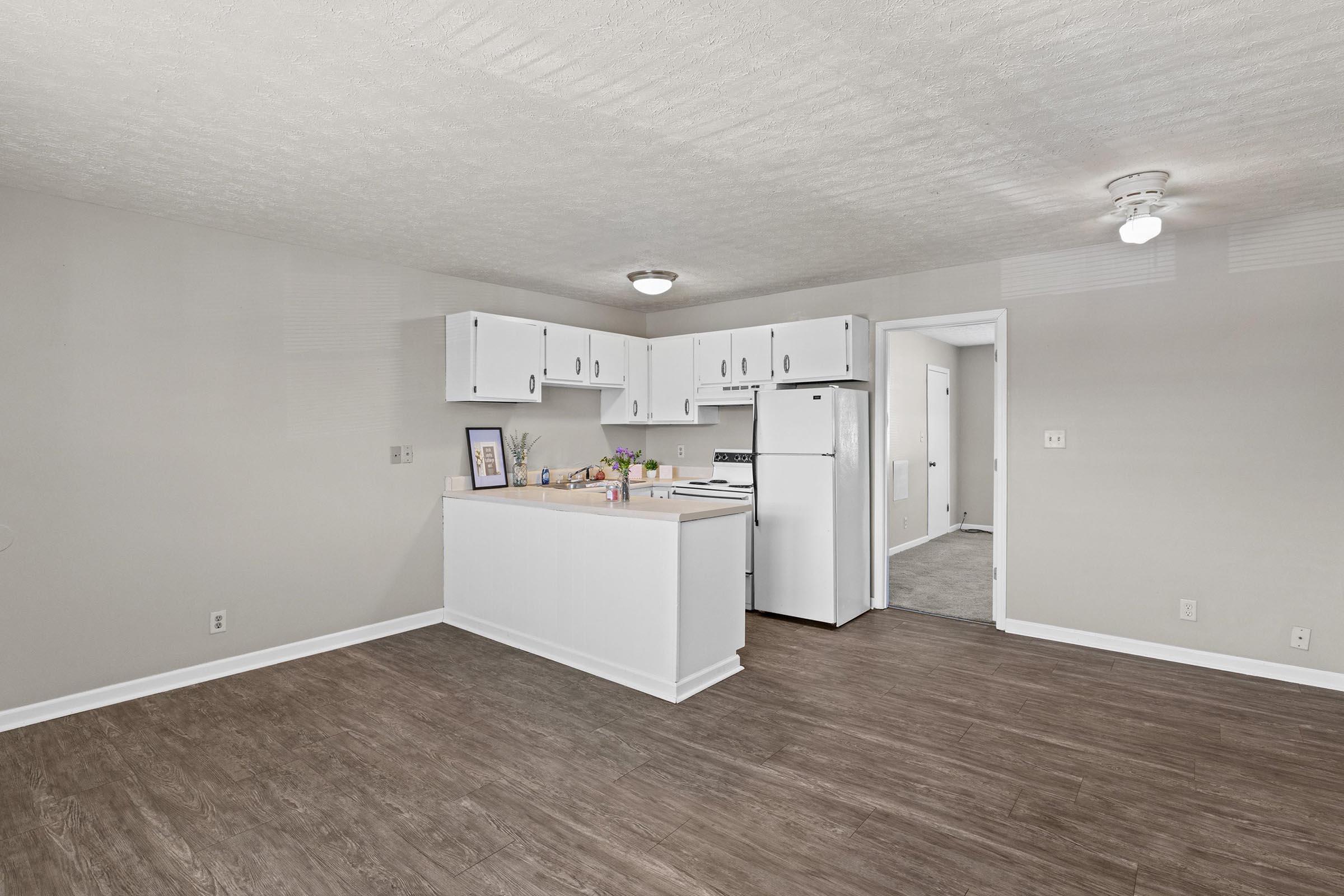 A spacious, modern kitchen in an apartment featuring white cabinetry, a refrigerator, and a stove. The area has a light-colored, textured ceiling, dark wood-like flooring, and a neutral wall color. A doorway leads to another room, creating an open and inviting atmosphere.