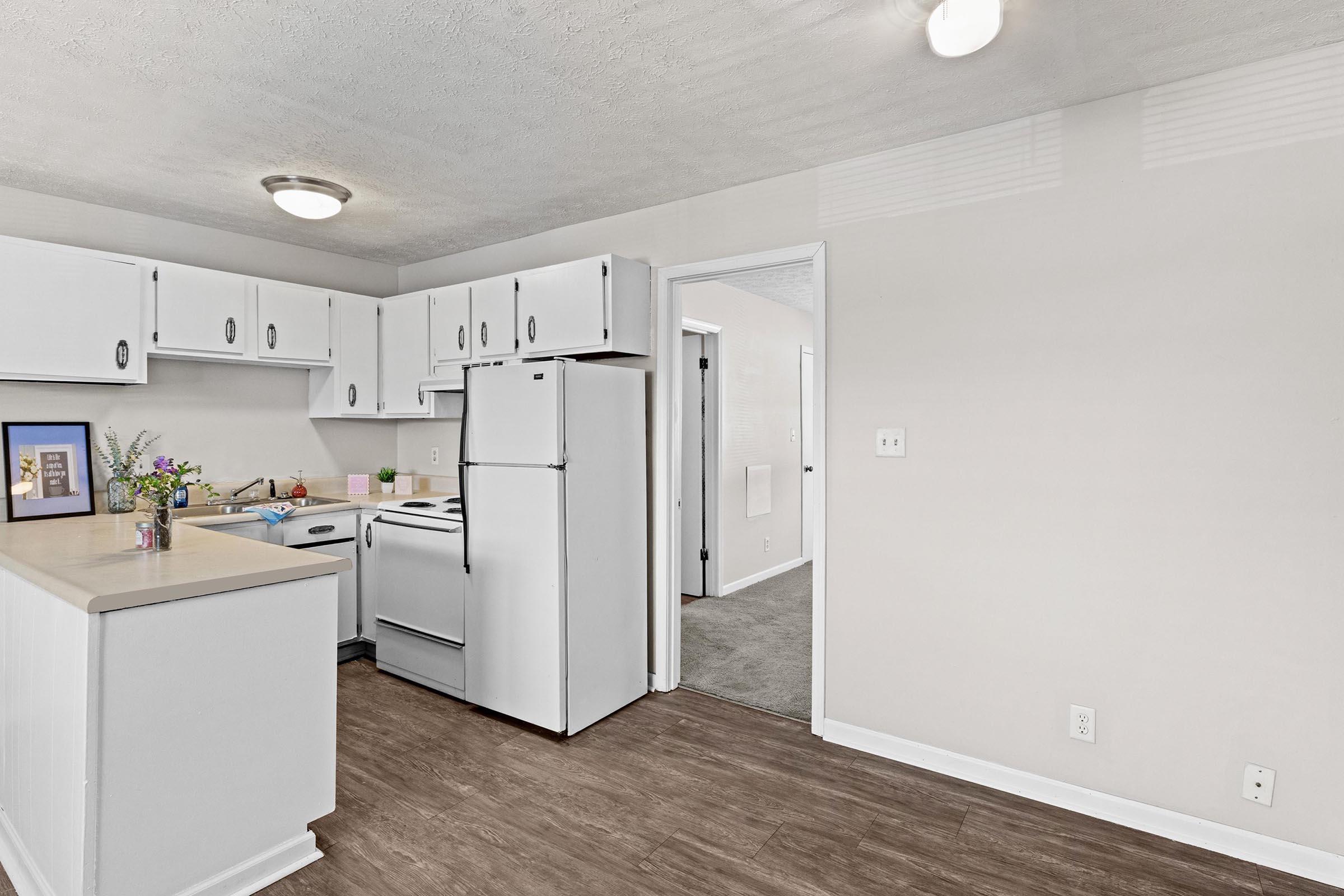 A modern kitchen featuring white cabinets, a refrigerator, and a stove. The countertop is clear except for a small vase of flowers. A doorway leads to another room, and the floor has a wooden appearance. Natural light enters the space, enhancing the bright and tidy atmosphere.