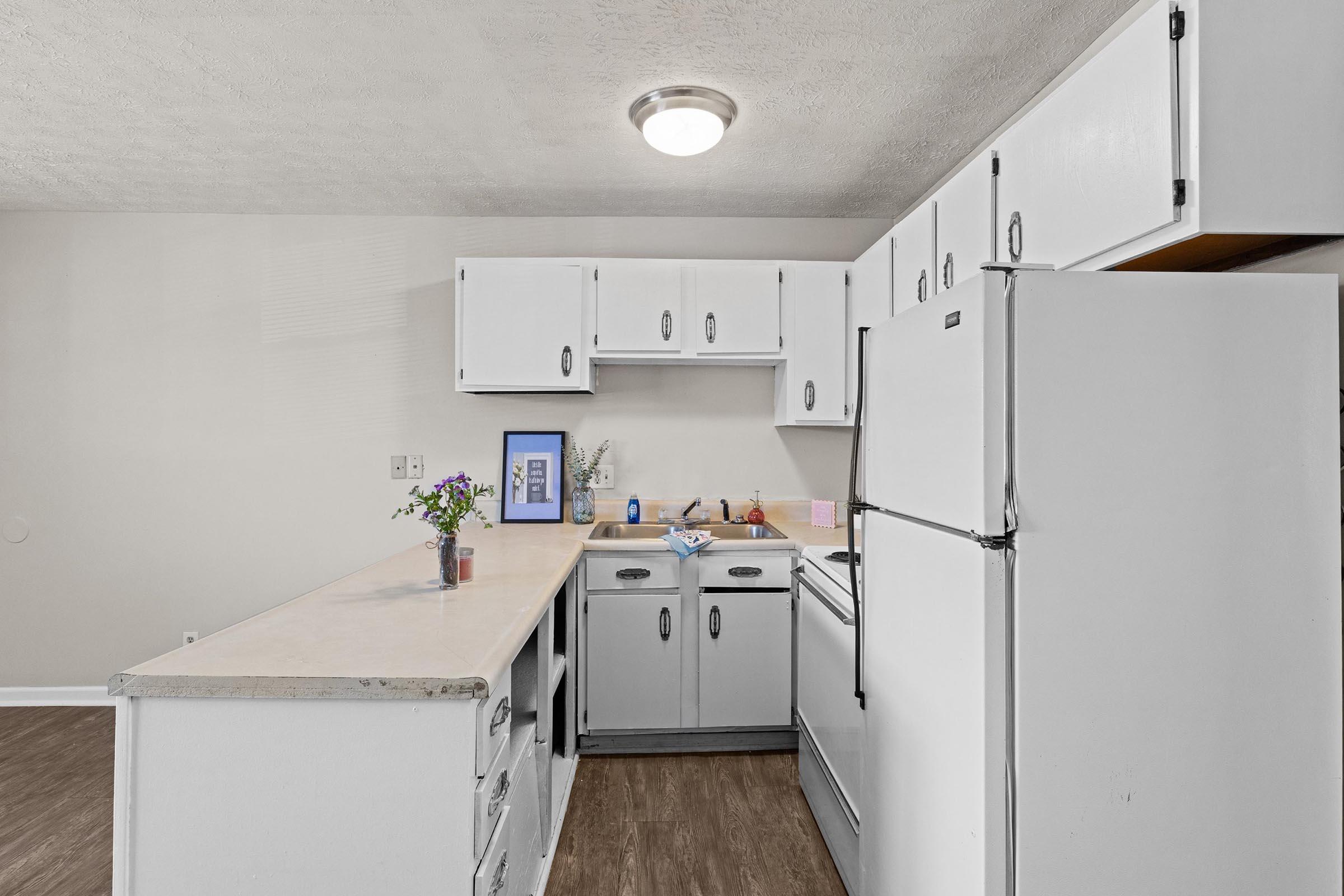 A bright, modern kitchen featuring white cabinets, a white refrigerator, and a countertop. A small vase with flowers is on the counter, alongside various kitchen items. The floor is a dark wood finish, and a ceiling light provides illumination. The overall atmosphere is clean and inviting.