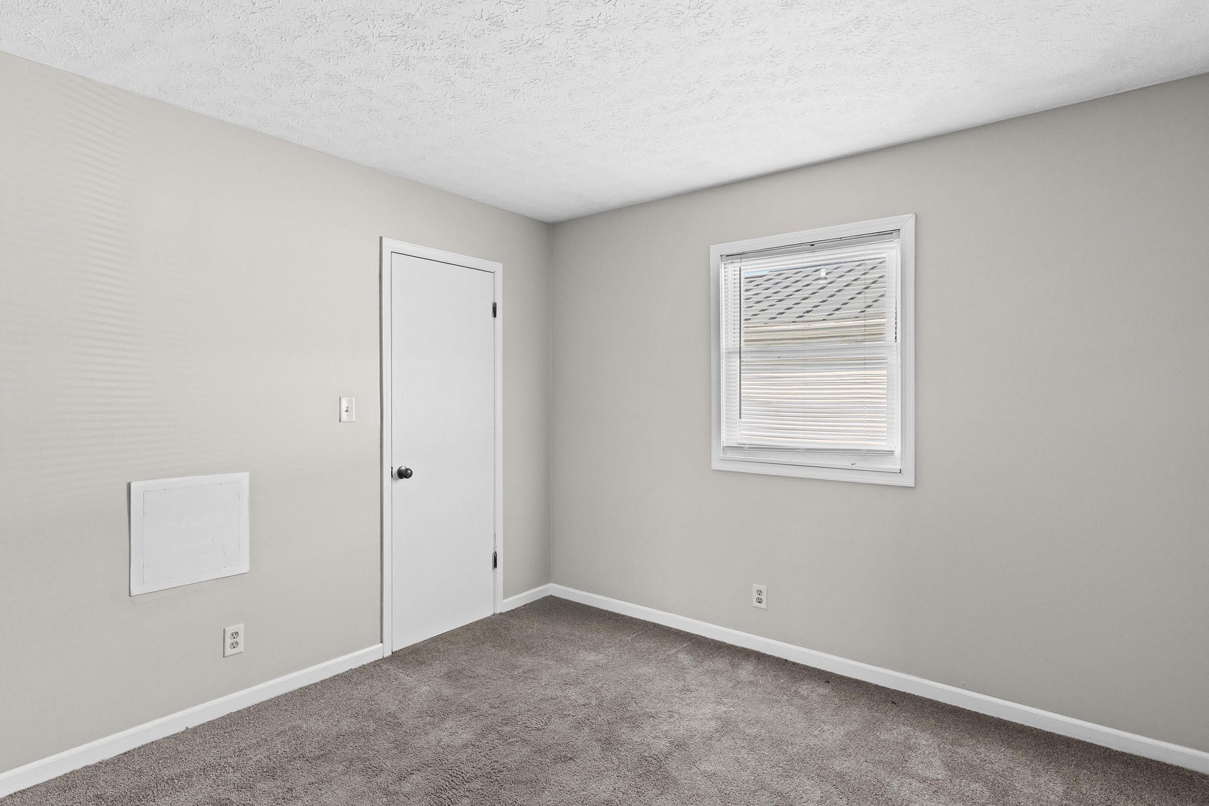 Empty room with light gray walls, a window with white blinds, a white door, and tan carpet. A small wall panel is visible on the left side of the room.
