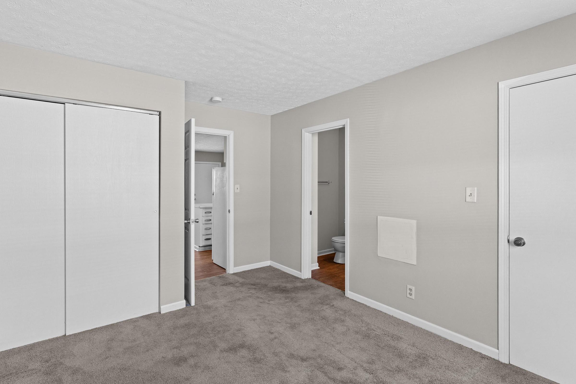 A view of a light-colored room featuring beige carpet, white walls, and doors leading to other areas. One door is open, revealing a bathroom, while another door leads to a closet. The room has a bright and neutral aesthetic, suitable for various uses.