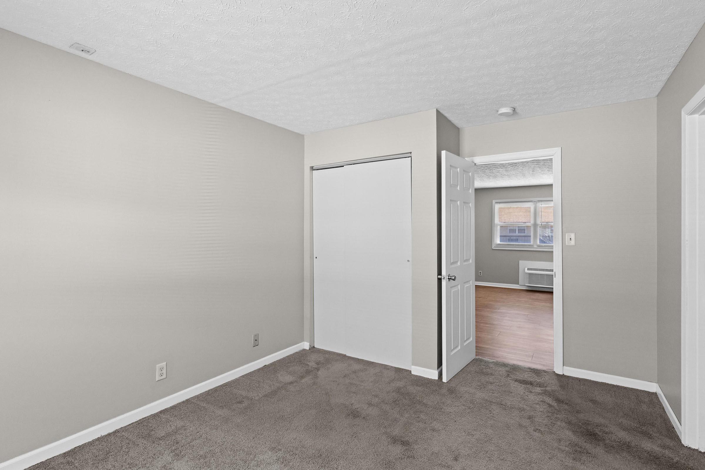 A spacious, empty room with light beige walls and carpeted flooring. The room features a white closet door, an open doorway leading to another room, and a window. Natural light illuminates the space, highlighting its neutral colors and clean lines.