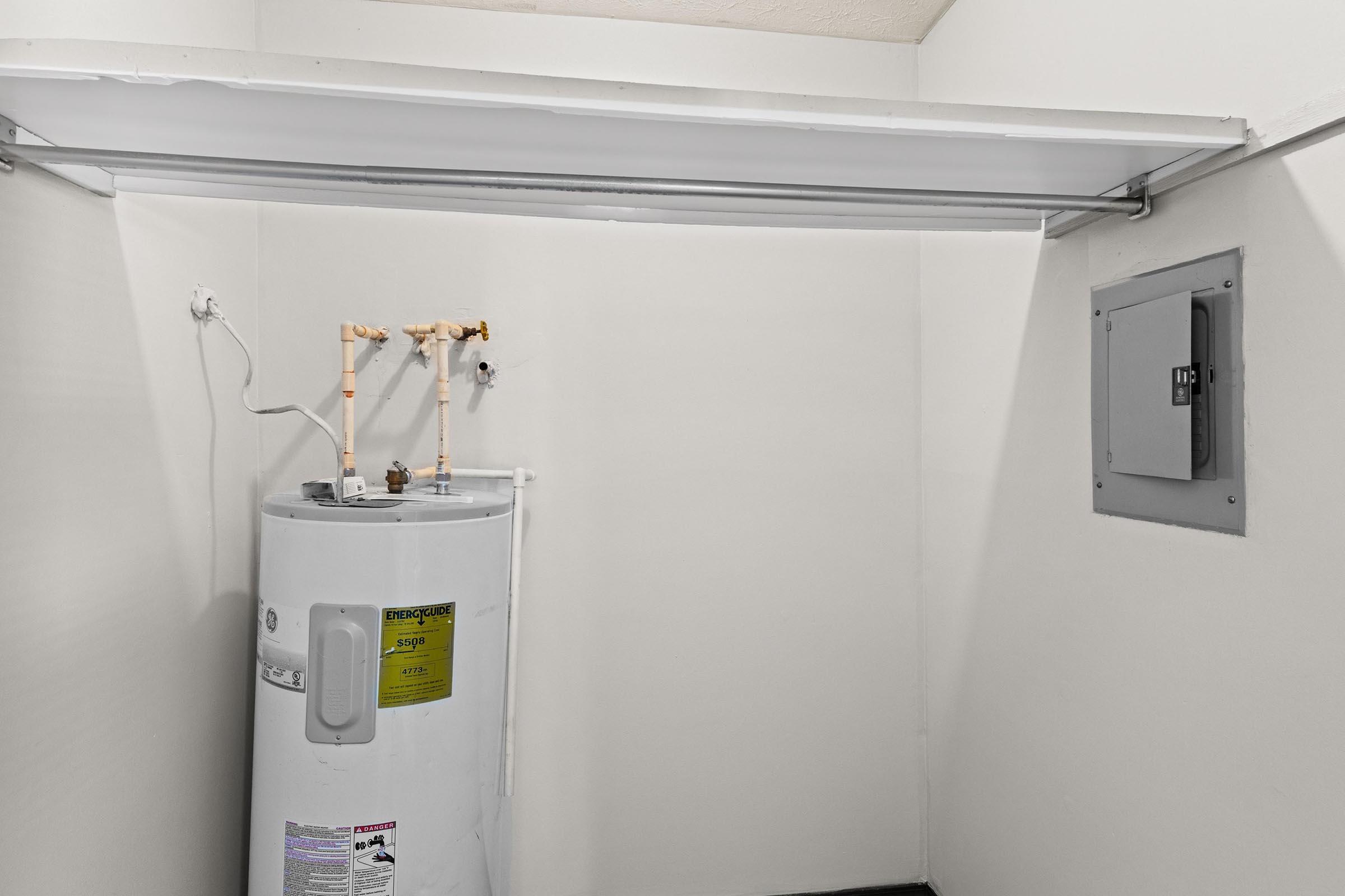 A clean, empty laundry room featuring a white hot water heater with copper pipes and a control panel on the wall. The walls are painted white, and there is a minimalist appearance with no additional furniture or items in the space.