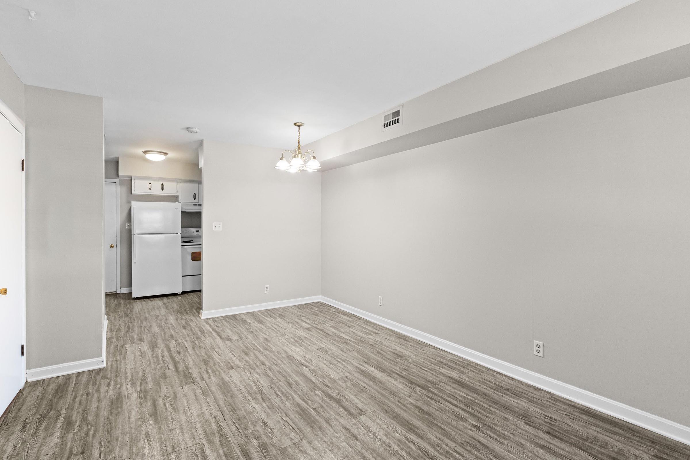 An empty, well-lit room with light gray walls and a wooden floor. There's a hanging light fixture with three bulbs. In the background, a kitchen area is visible, featuring white appliances including a refrigerator and an oven. The space is clean and ready for furnishing.