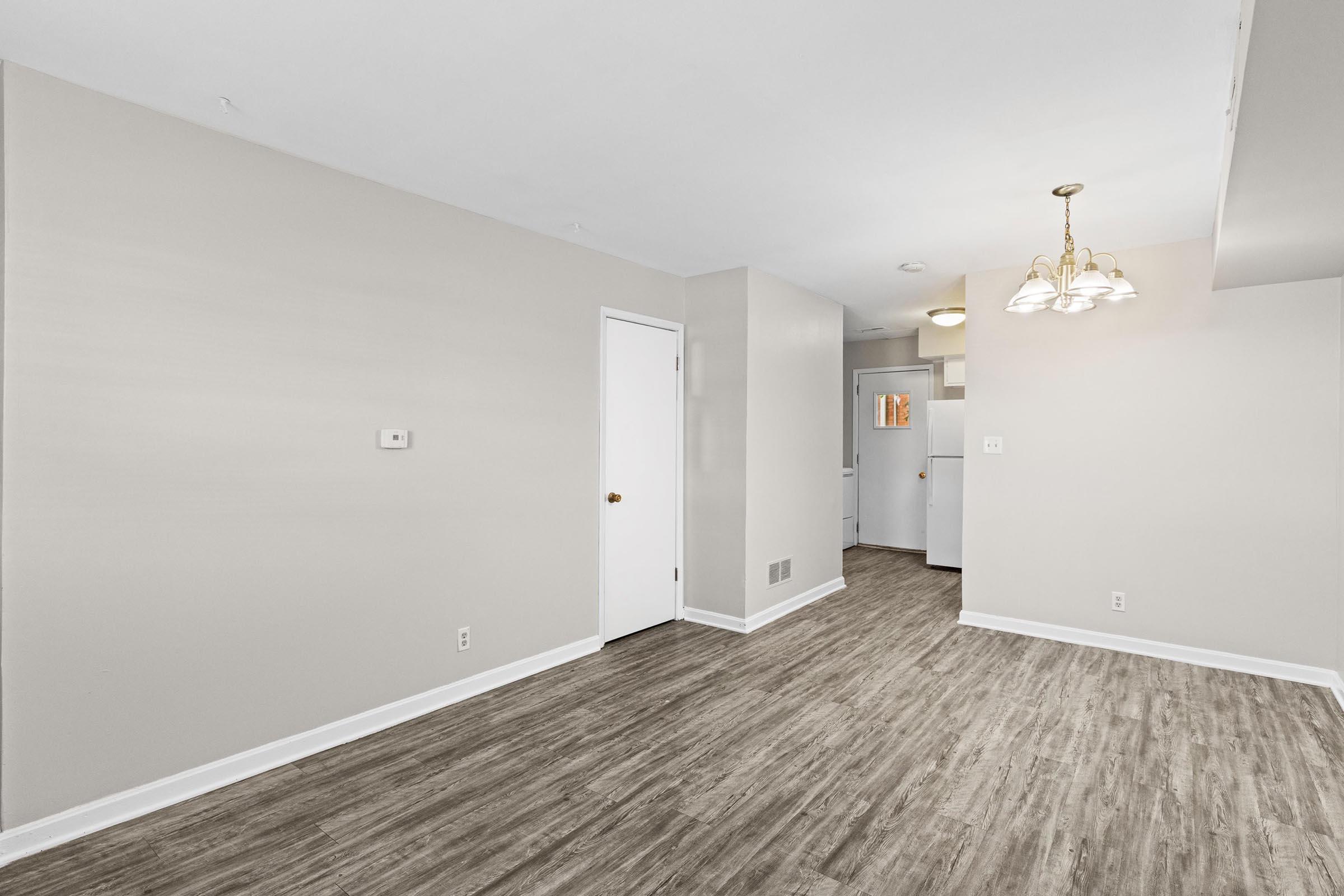 A spacious, empty room with light-colored walls and a wood-style floor. On the left, there is a white door, and in the background, a well-lit kitchen area is visible. A light fixture with three bulbs hangs from the ceiling, creating a welcoming ambiance.