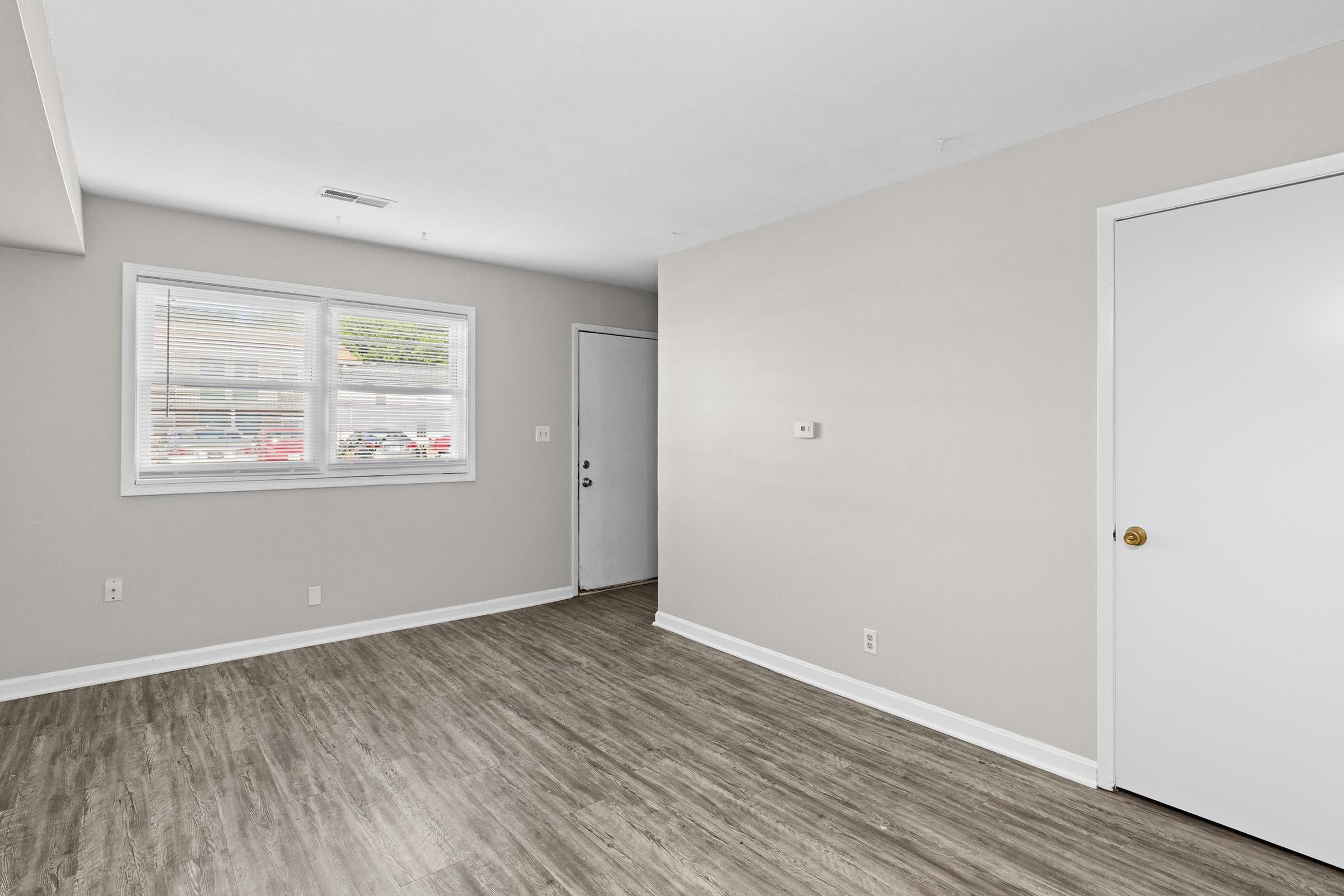 An empty room featuring a light-colored wall, a door on the right, a window with blinds on the left, and a floor with a wood-like finish. The space is well-lit with natural light coming through the window.