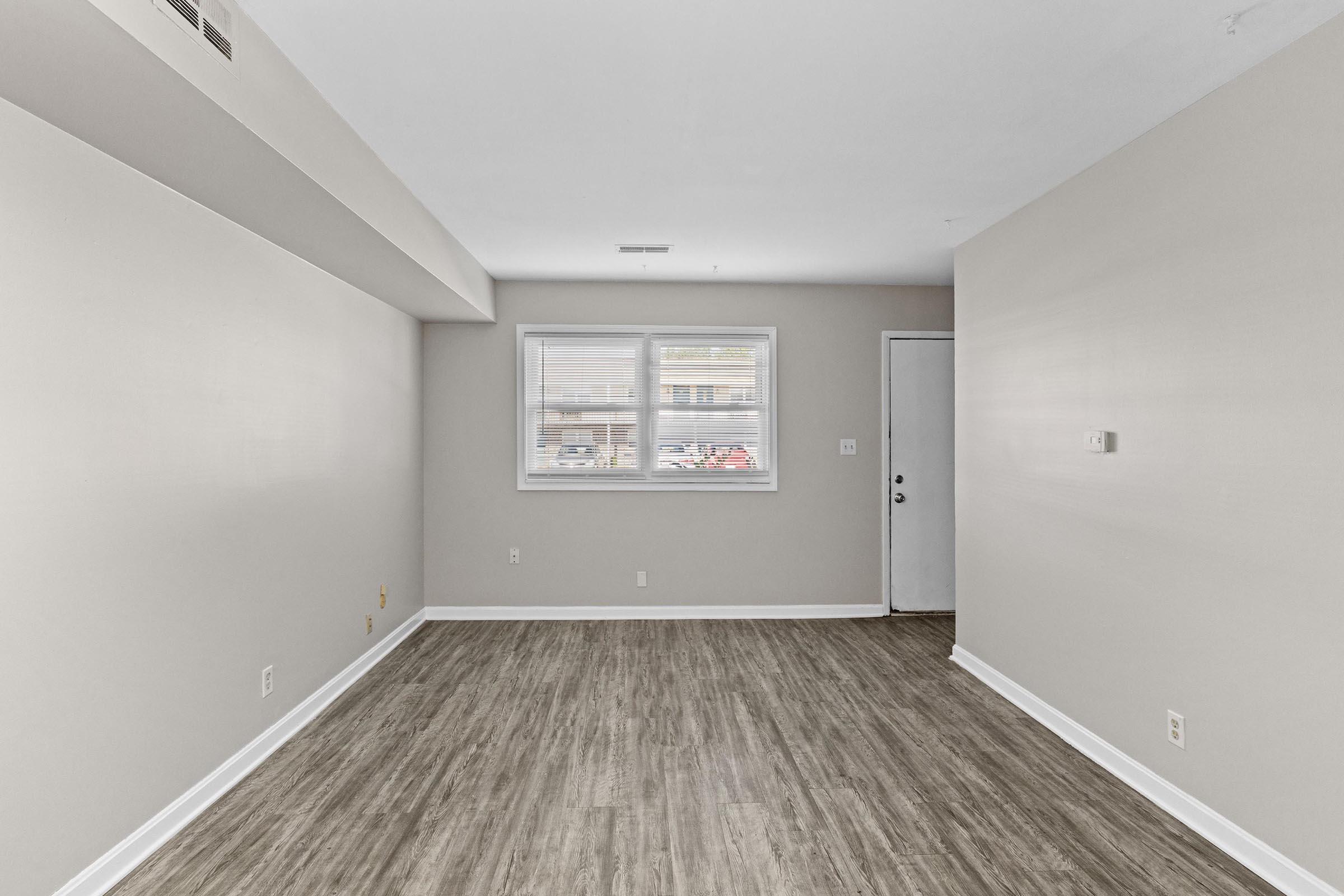 A spacious, empty room with light gray walls and a wooden floor. It features two windows allowing natural light to enter, facing an exterior view. A single door is visible on the right side, suggesting access to another area. The room is clean and ready for furnishings.