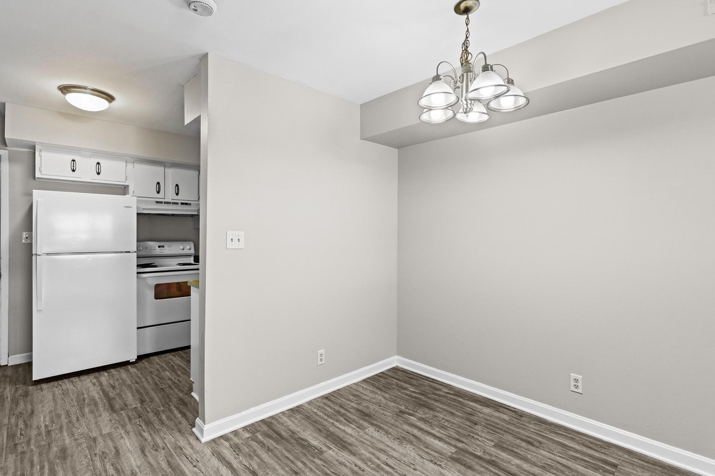 A bright, empty room with light-colored walls and a modern chandelier. To the left, there's a doorway leading to a kitchen featuring a white refrigerator and stove. The floor has a wood-like finish, adding warmth to the space. Natural light creates a welcoming atmosphere.