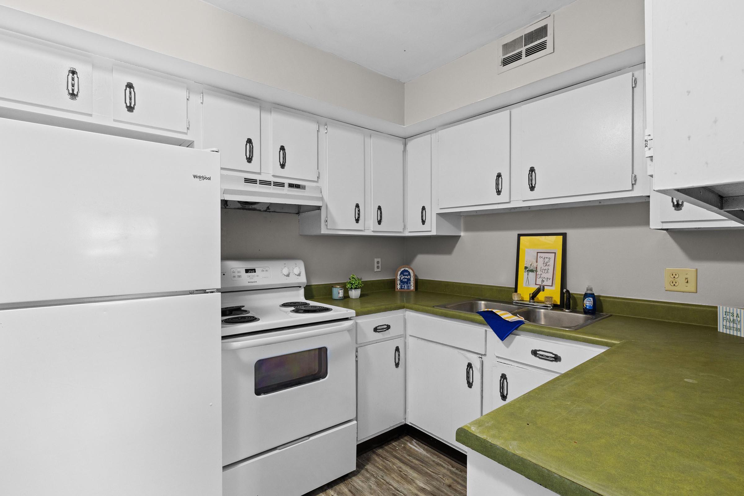 A kitchen featuring a white refrigerator, an electric stove with an oven, and white cabinets. The countertops are green, and there's a stainless steel sink. A blue kitchen towel and a small plant are on the counter, along with a framed picture on the wall. The overall design is simple and functional.