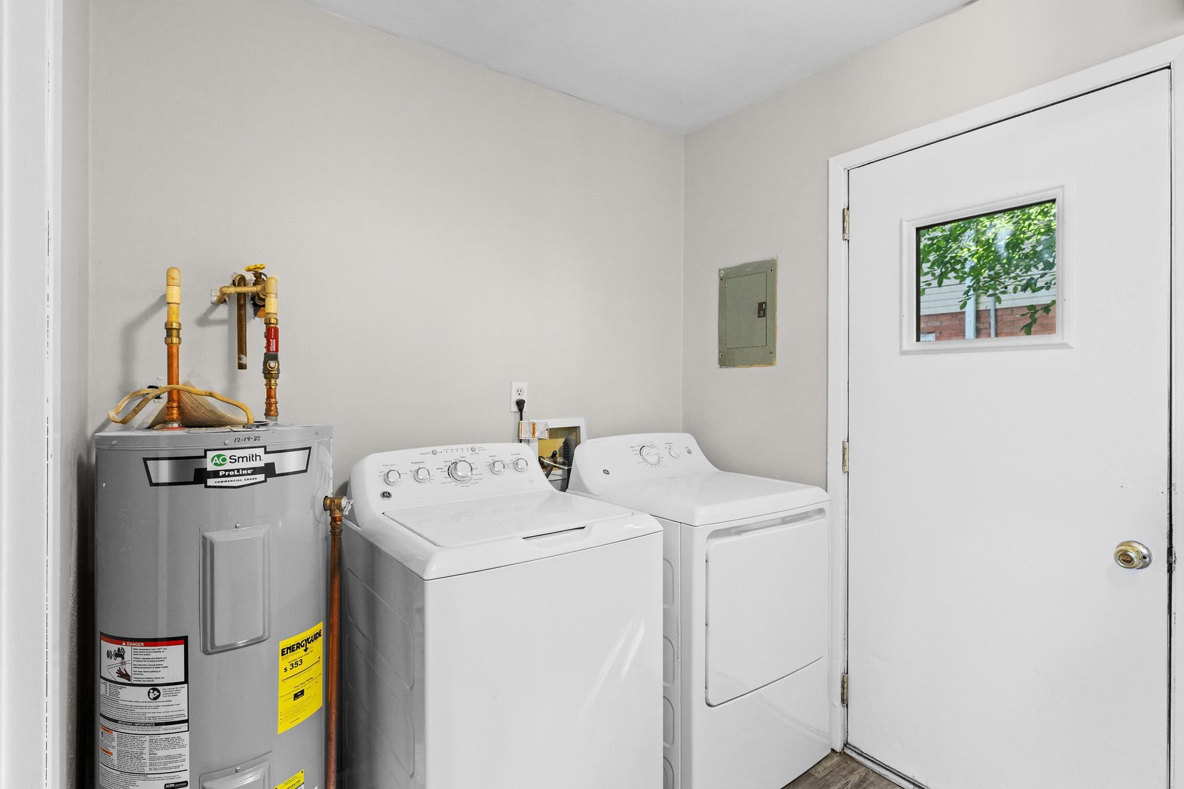 A laundry room featuring a gray water heater, a white washing machine, and a white dryer. The room has light beige walls and a door with a window, providing natural light. A small electrical panel is visible on the wall next to the appliances.