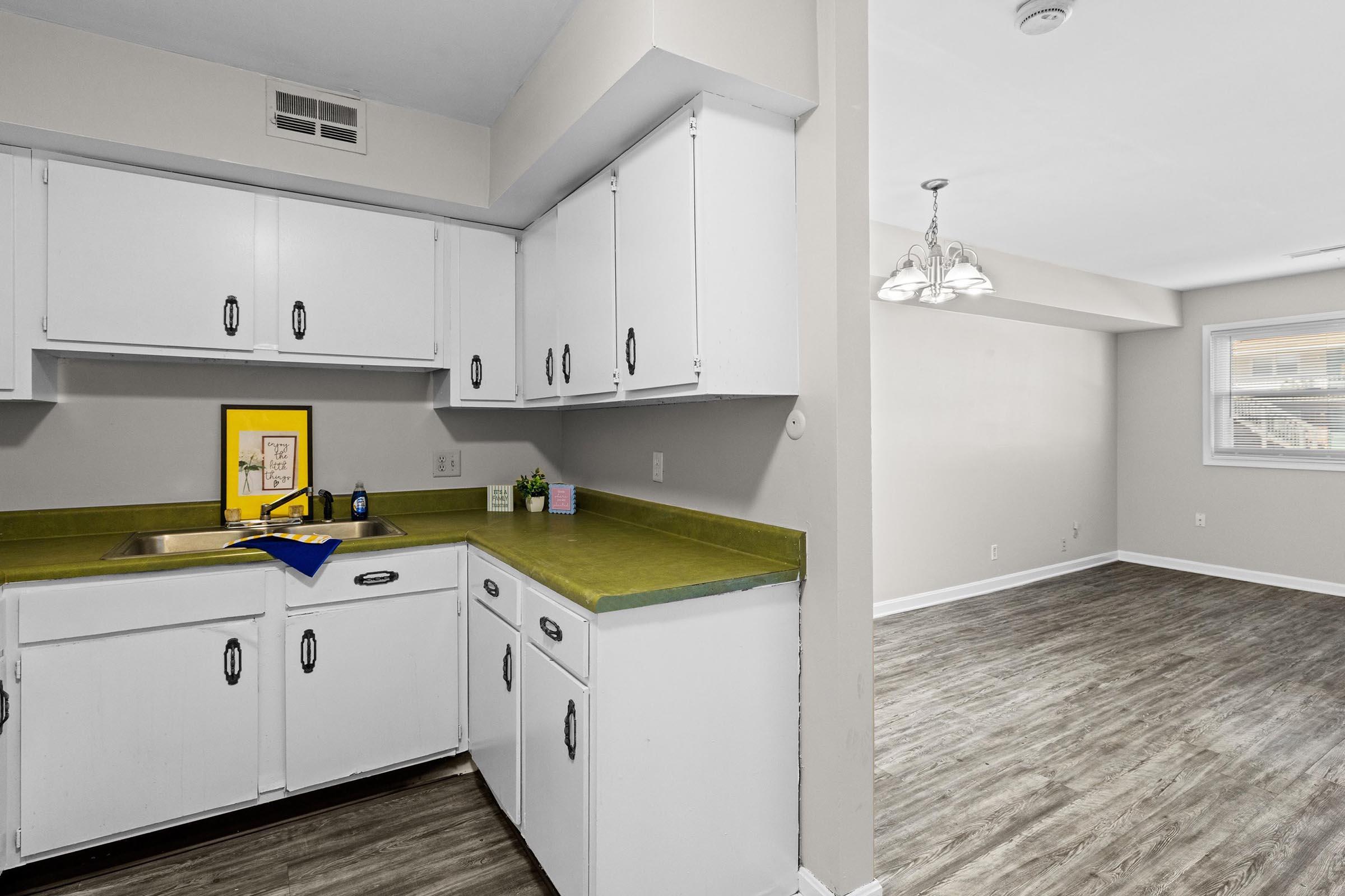 A modern kitchen with white cabinets and a green countertop, featuring a sink and a framed picture on the counter. The room opens into a spacious living area with light-colored walls and a large window, showcasing wood-like flooring. A dining area is visible with a chandelier hanging above.