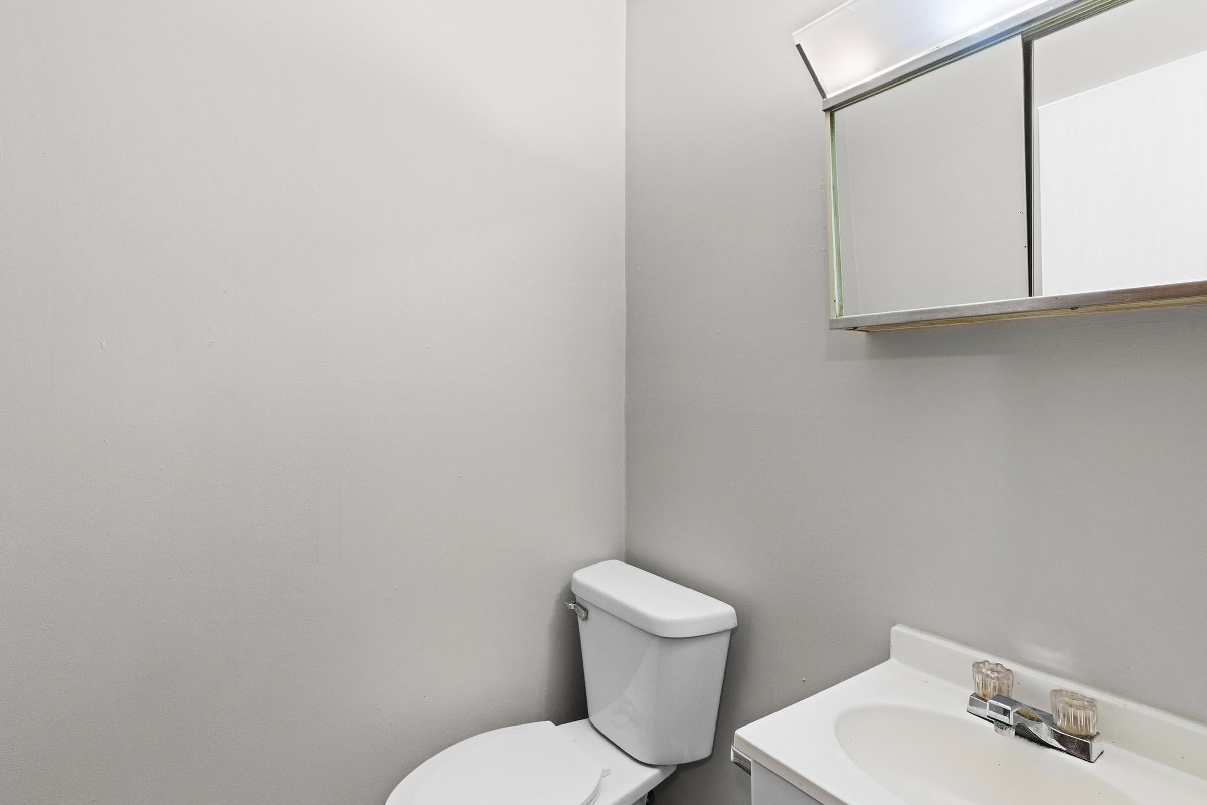 A small, minimalistic bathroom featuring a white toilet, a sink with a faucet, and a mirror above the sink. The walls are painted a light gray, creating a neutral backdrop. The space appears clean and uncluttered.