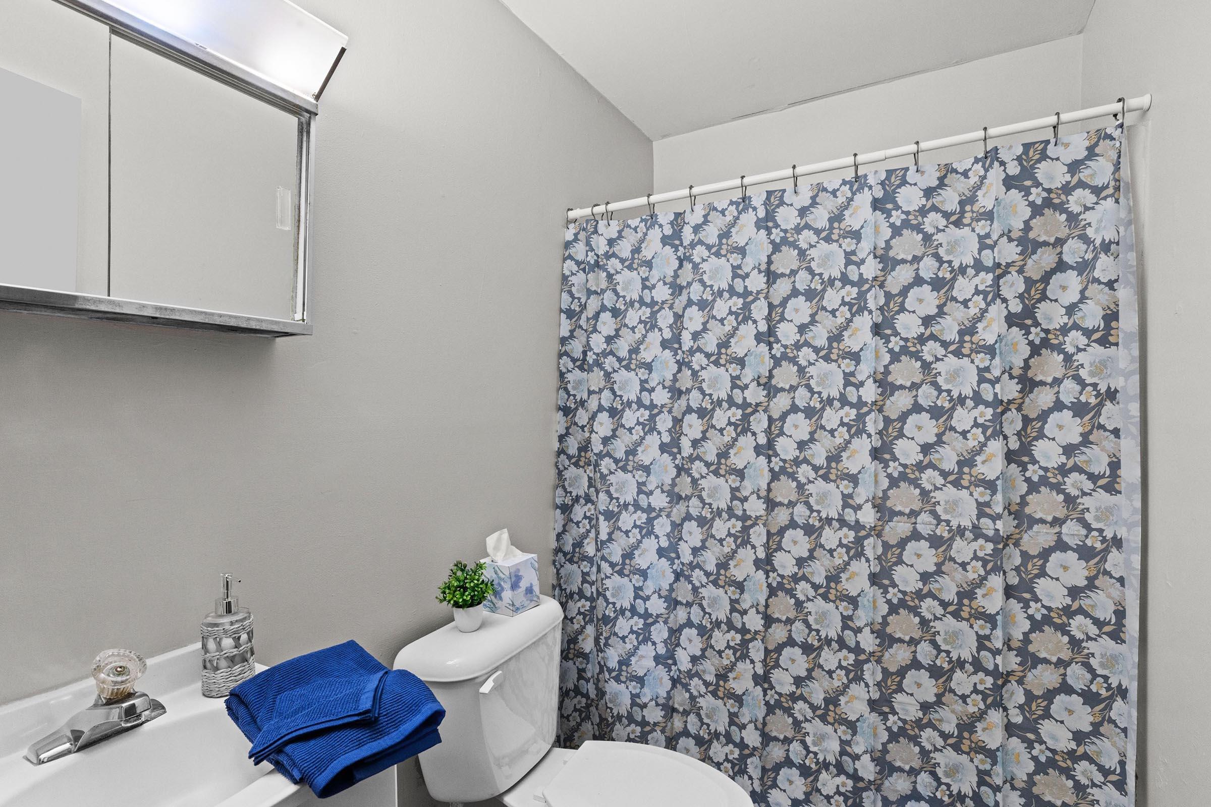Brightly lit bathroom featuring a white toilet and sink. A shower curtain adorned with a floral pattern in shades of blue covers the bathtub. A small plant and tissue box sit atop the toilet tank, alongside neatly folded blue towels. A mirror is mounted above the sink, reflecting the room's light.