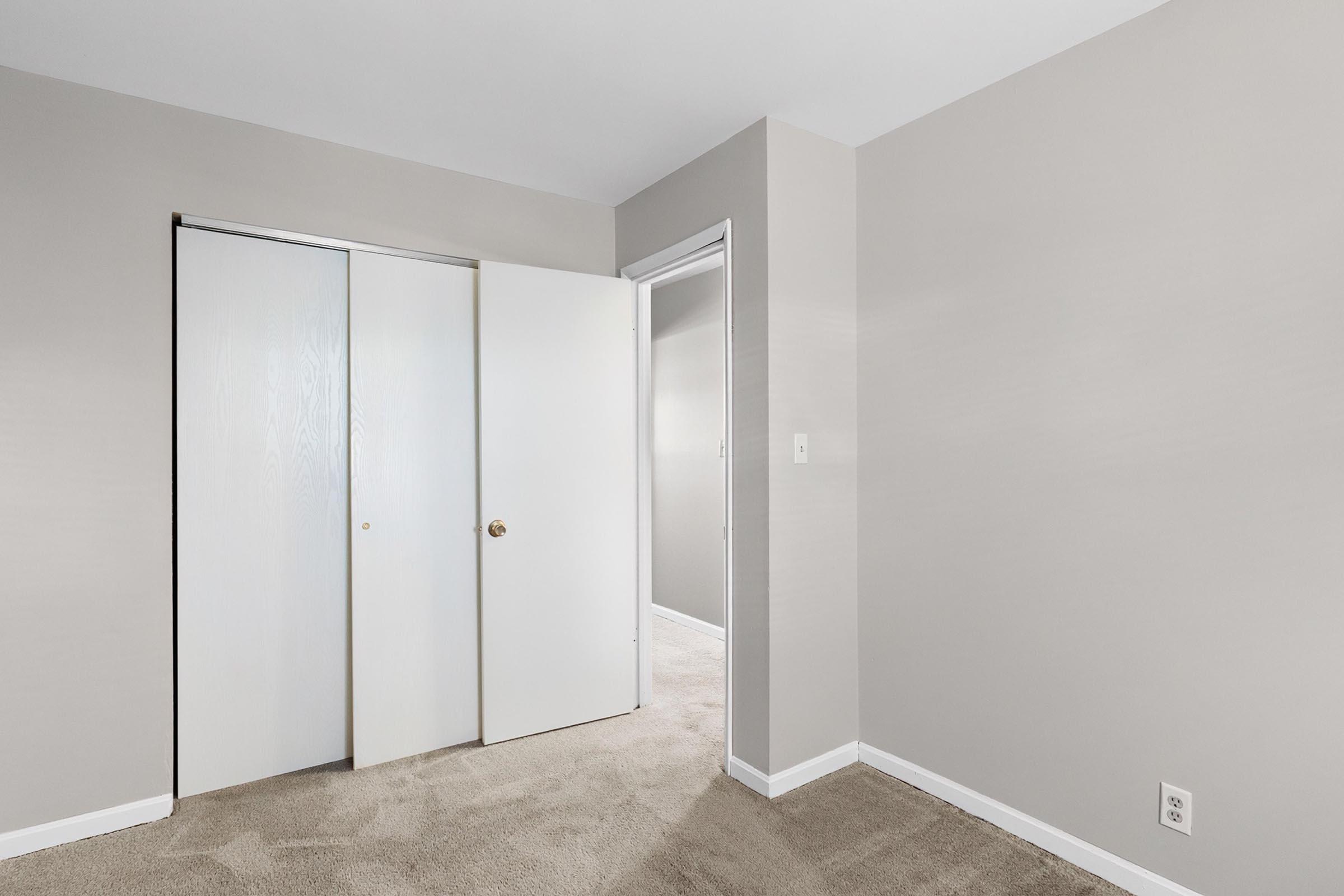 A well-lit interior room featuring light gray walls, a beige carpeted floor, and a pair of white sliding closet doors. There is a doorway leading to another room, with a simple, clean design. The space appears tidy and spacious, ideal for use as a bedroom or office.