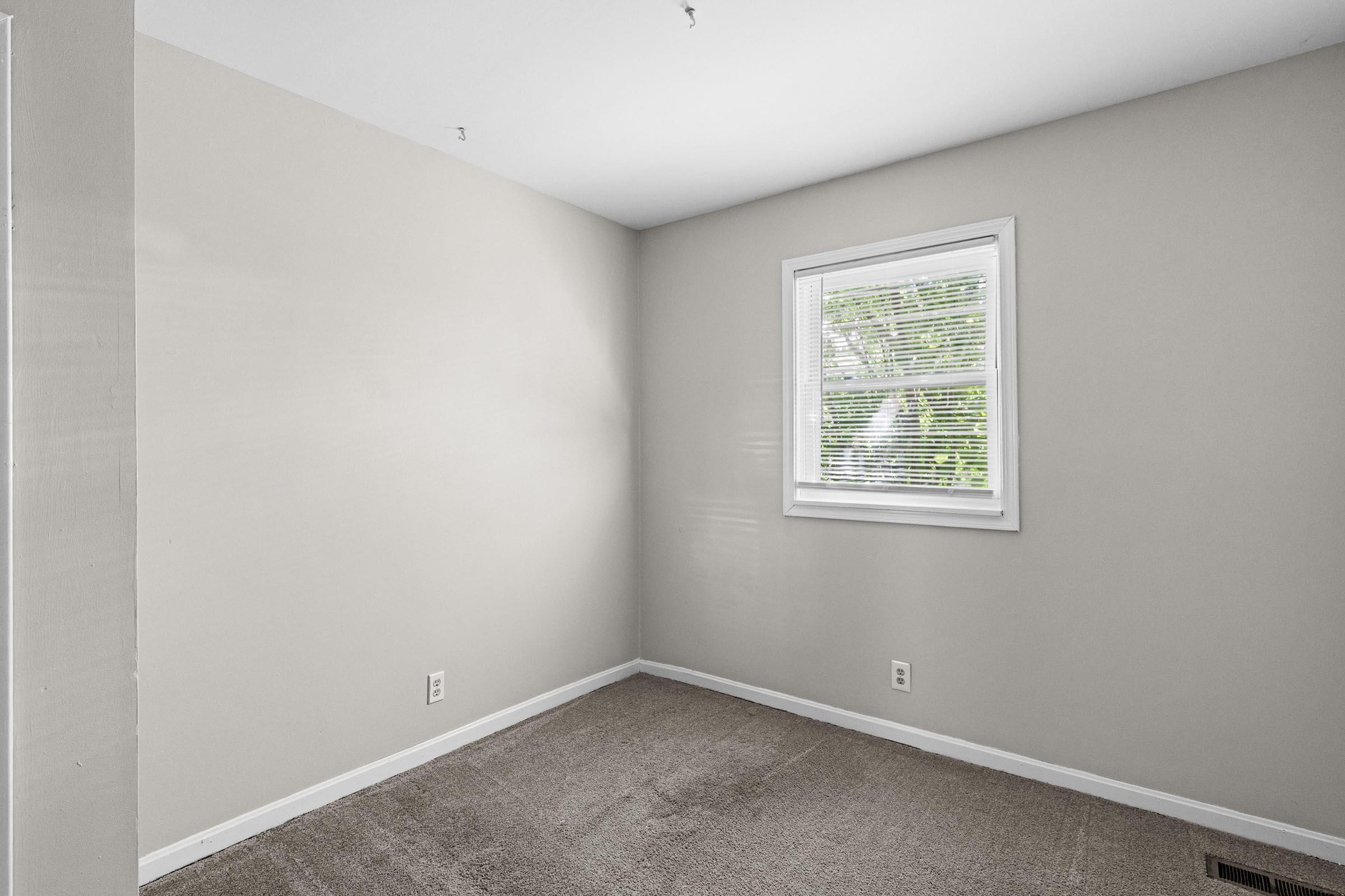 A small, empty room with light beige walls and a carpeted floor. There is a window with white blinds on one wall, allowing natural light to enter, and no furniture or decorations present, creating a clean and uncluttered appearance.