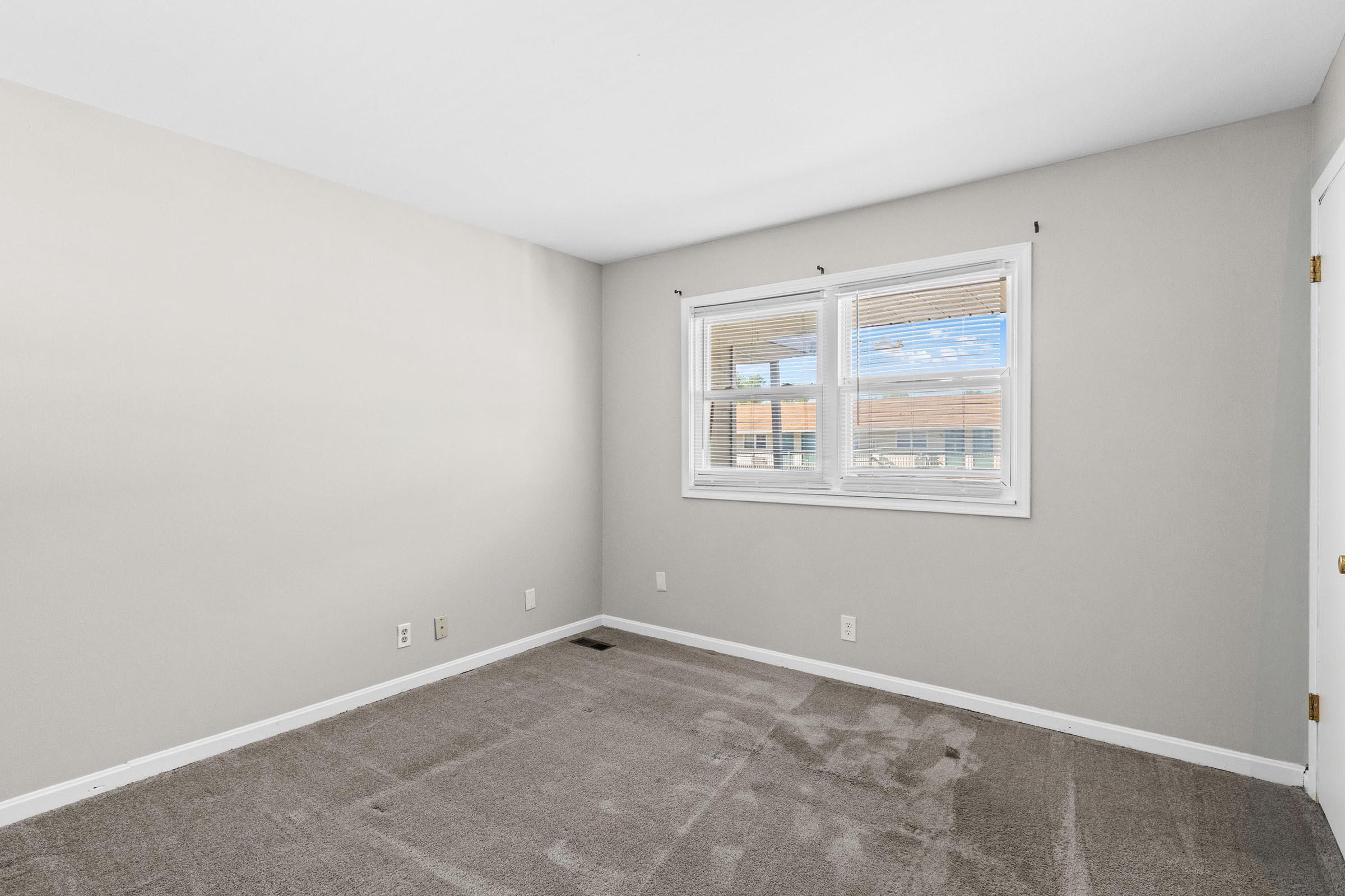Empty room with light gray walls and a carpeted floor. Two windows with white trim allow natural light to enter. No furniture or decorations are present, creating a spacious and minimalistic atmosphere. A door is visible on the right side of the image.