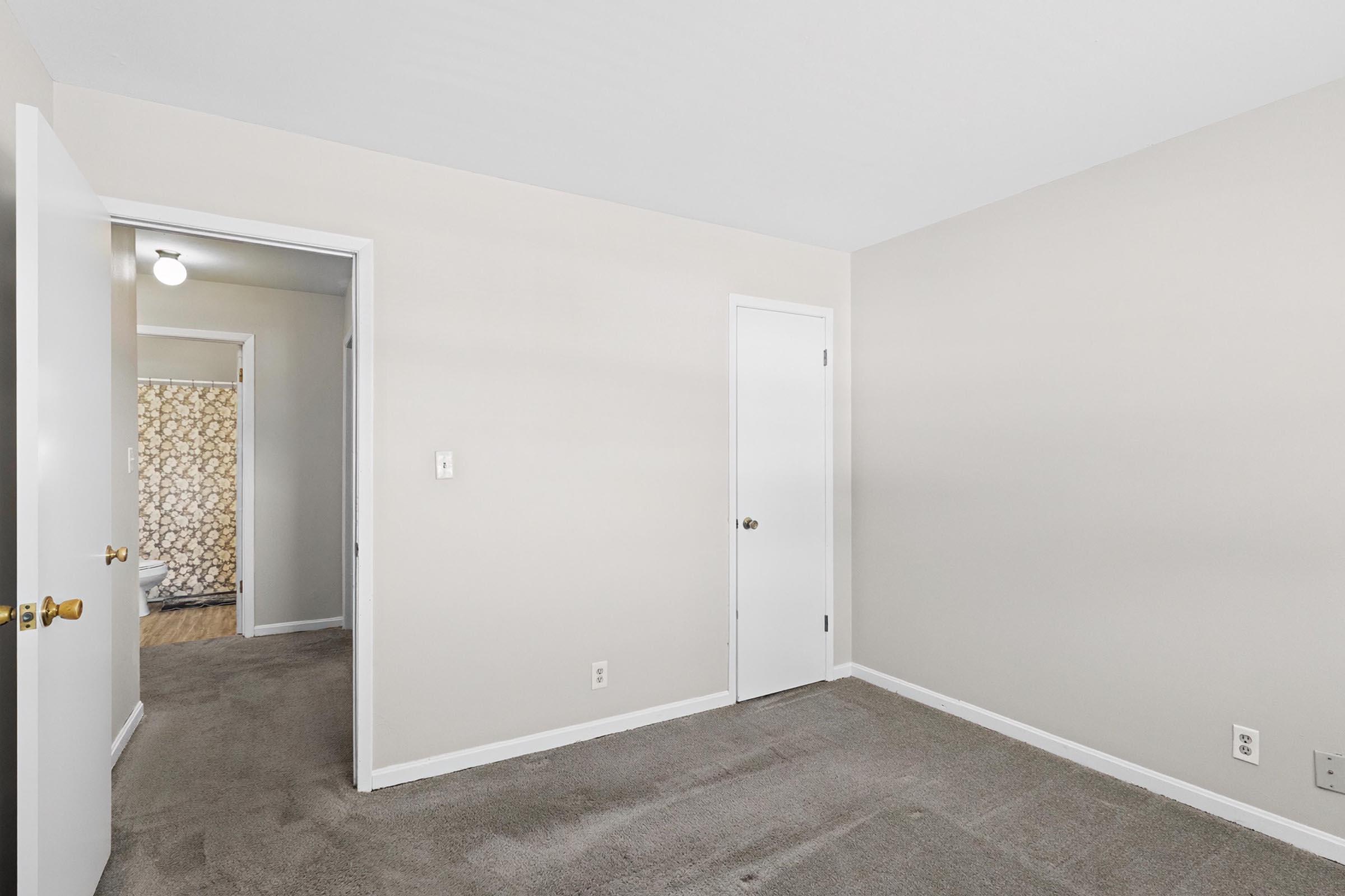 A vacant room with light beige walls and carpet. There are two doors: one leads to a hallway and the other to a bathroom, which is partially visible. The room is well-lit from a ceiling light fixture, and the floor features a neutral carpet texture.