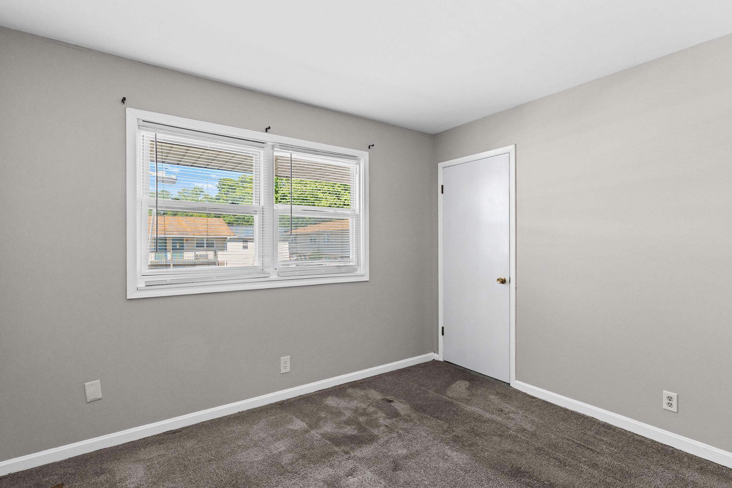 Empty room with light gray walls and brown carpet. Two windows with blinds allow natural light, and a white door is on one wall. The overall space appears clean and minimalistic, suitable for furnishing.