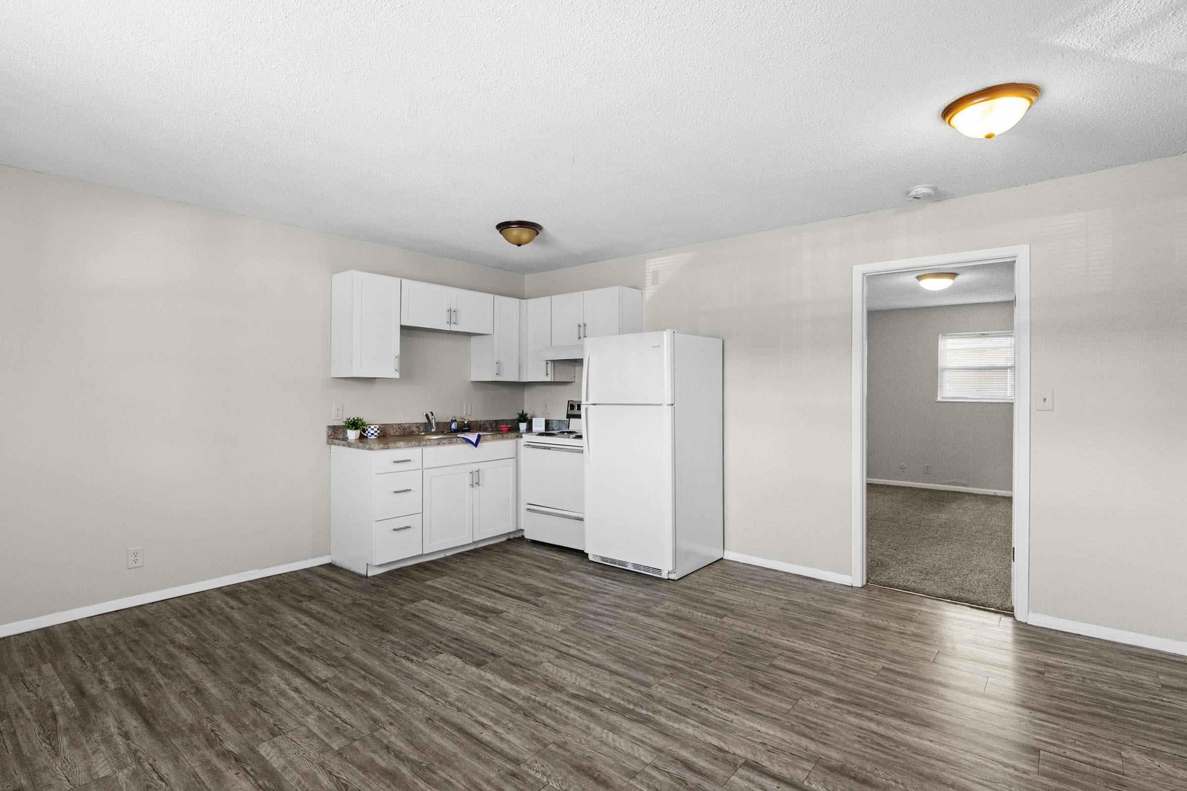A well-lit kitchen with white cabinets, a refrigerator, and a stove. The floor is hardwood, and there is a doorway leading to another room. The walls are painted light colors, creating a spacious feel in the area.