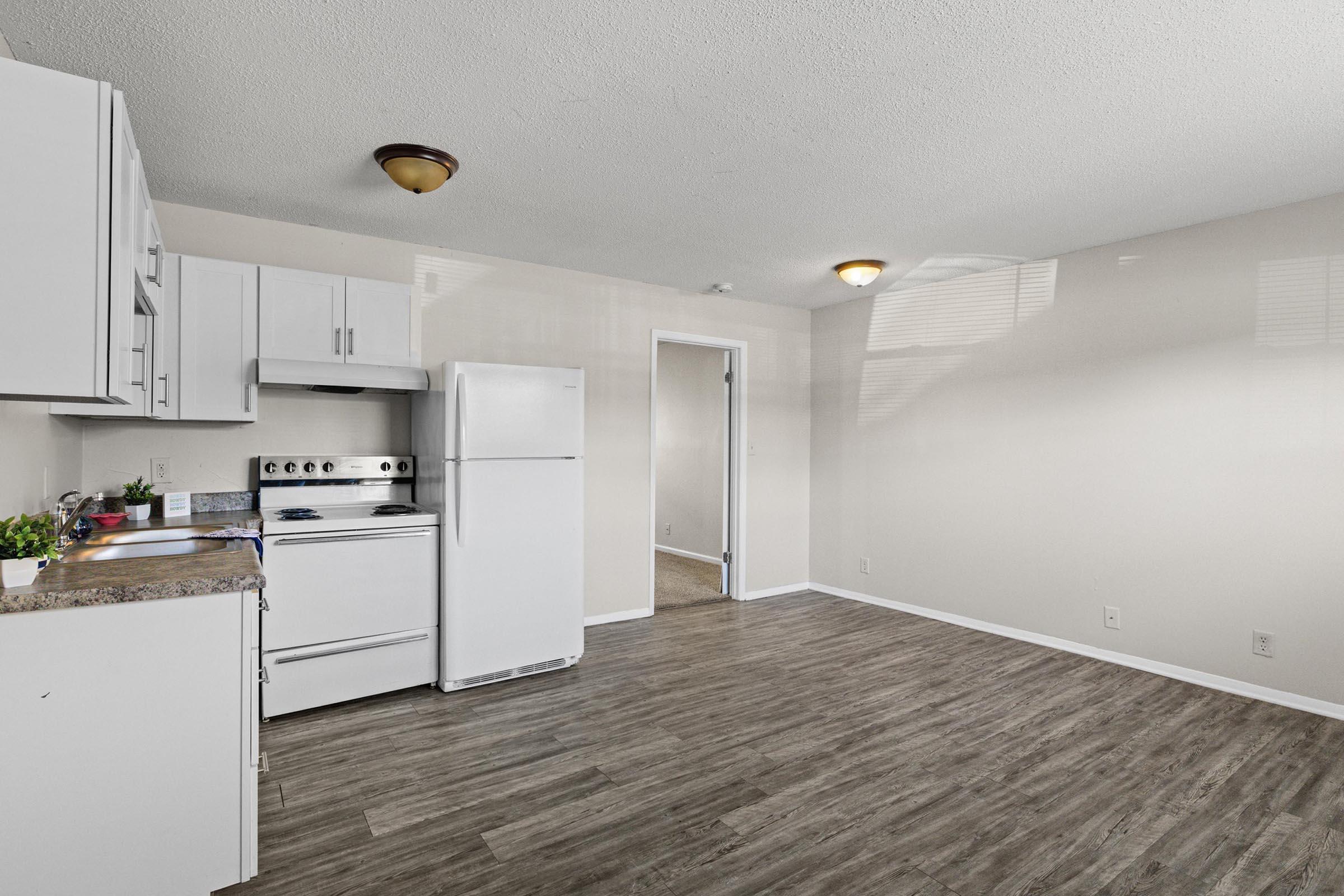 Modern kitchen with white cabinetry, stainless steel appliances (including a stove and refrigerator), and a countertop. The space features a light wood floor and neutral-colored walls. A doorway leads to another room, with natural light coming from a window. Simple and clean design, suitable for a living area.