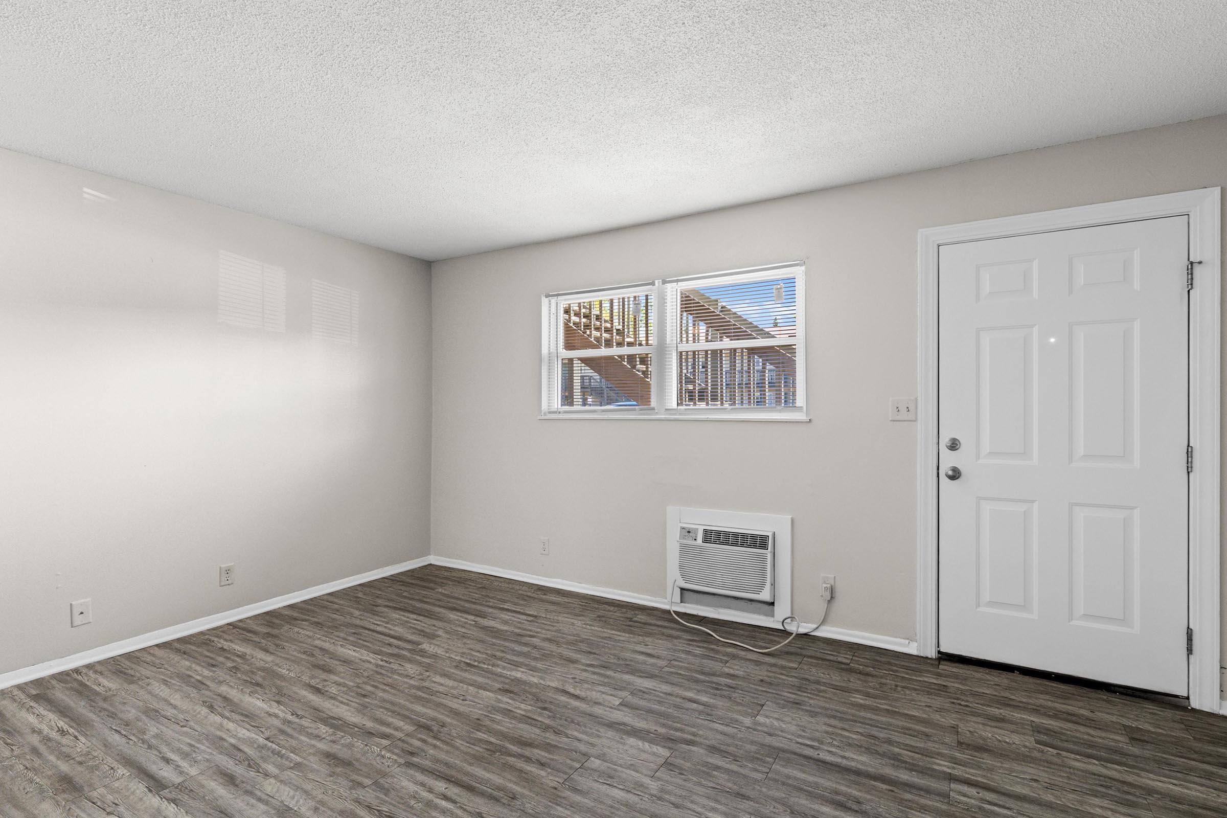 A small, empty room with light-colored walls and a wood-style floor. There is a white door on the right side, and a window on the opposite wall allowing natural light to enter. An air conditioning unit is installed below the window, and the space has a clean, minimalist appearance.
