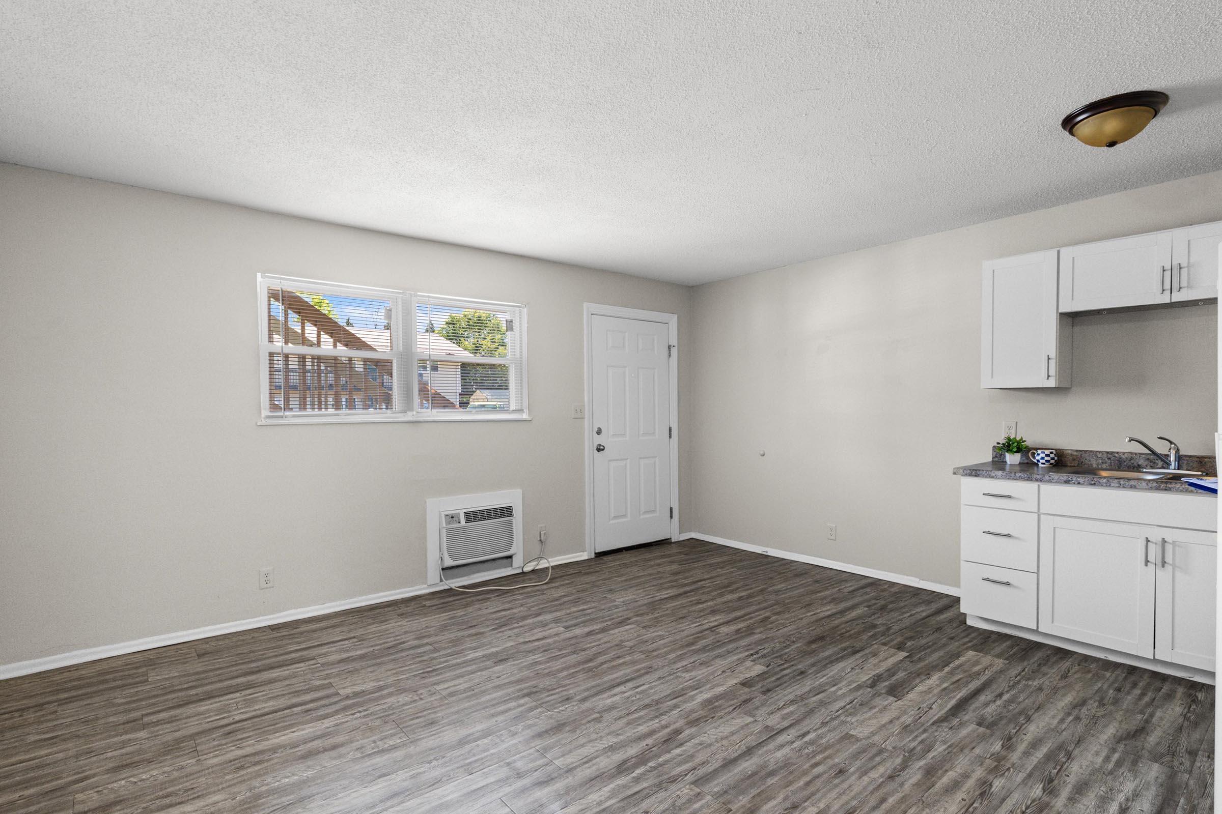 Empty room featuring a light-colored wall, a window with blinds, and a door. The flooring is a wood-like laminate. A small air conditioning unit is visible on the wall, and there is a kitchen area with white cabinets and a countertop along one wall. The overall space is bright and uncluttered.