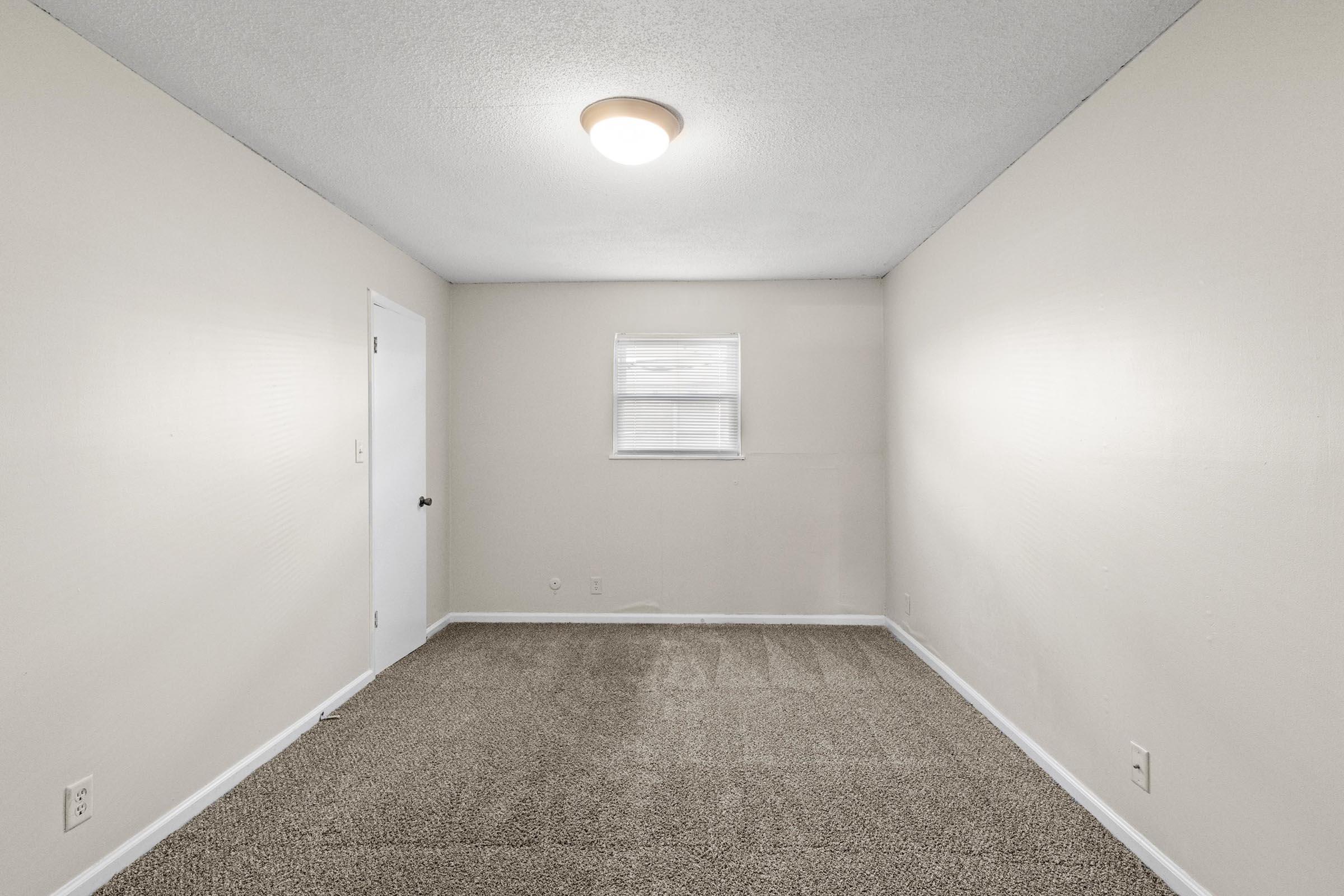 A blank room with light beige walls and a textured carpet. There's a single window with blinds on one wall and a closed white door on another wall. A ceiling light fixture illuminates the space, creating a neutral and simple aesthetic.