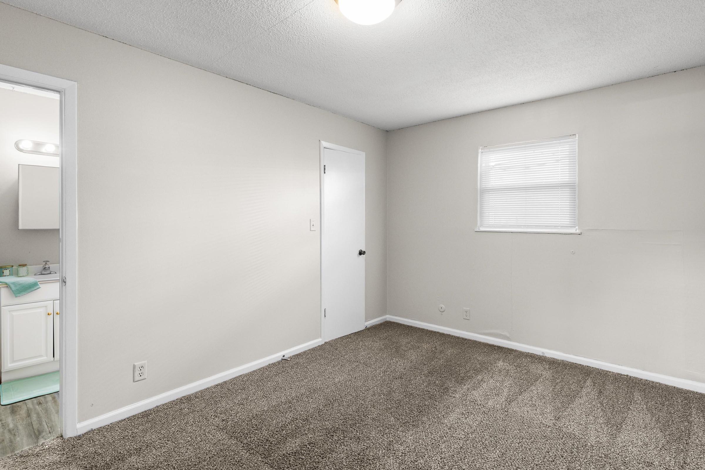 A sparsely furnished room with light beige walls and carpet. A white door leads to a bathroom visible in the background on the left. A small window allows natural light to enter. The room has a single ceiling light fixture and appears clean and well-maintained.