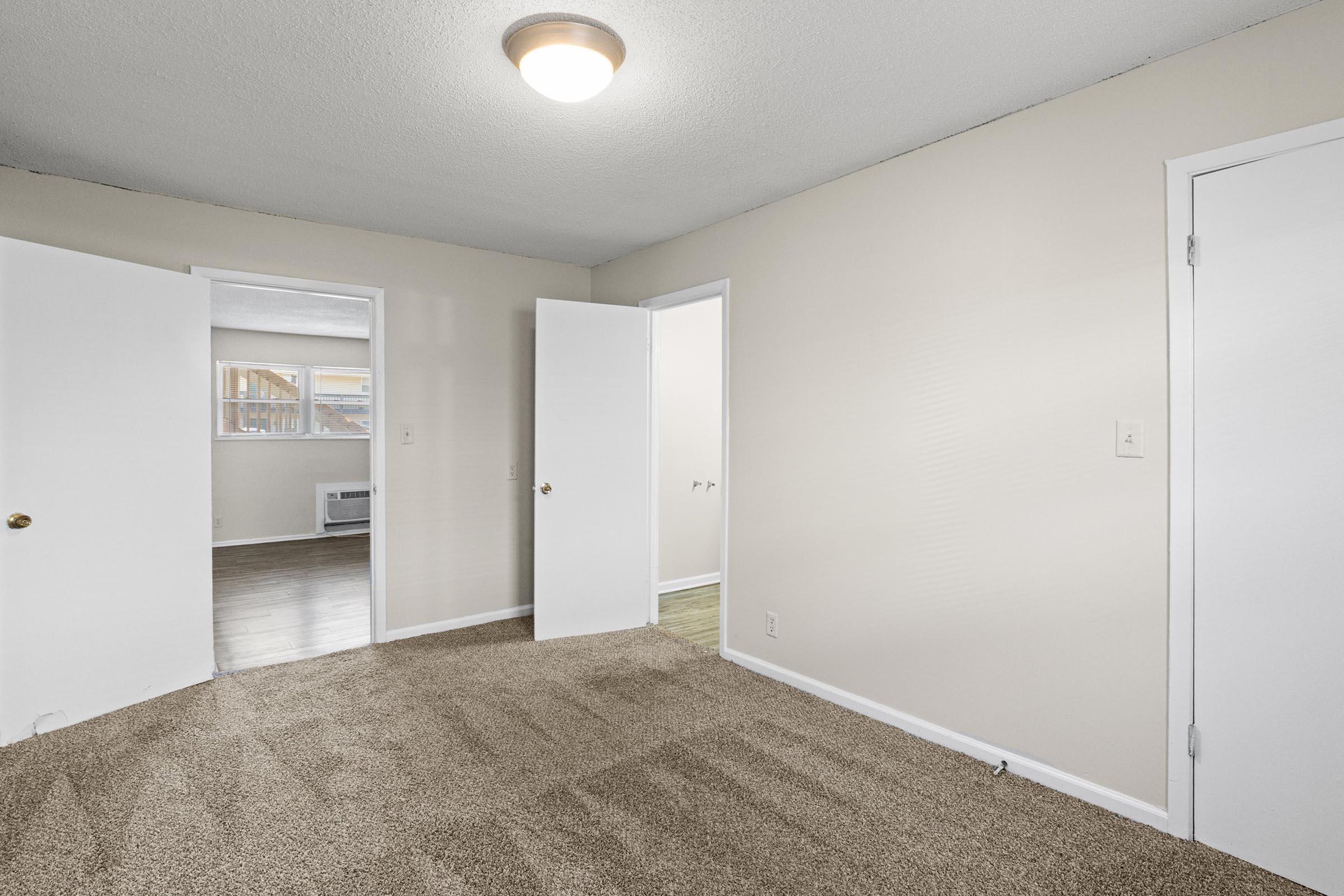 A light-colored room with beige carpet, featuring two open white doors leading to adjacent rooms. The walls are painted a soft beige, and there is a ceiling light fixture. A window is visible in the background, allowing natural light to enter the space.