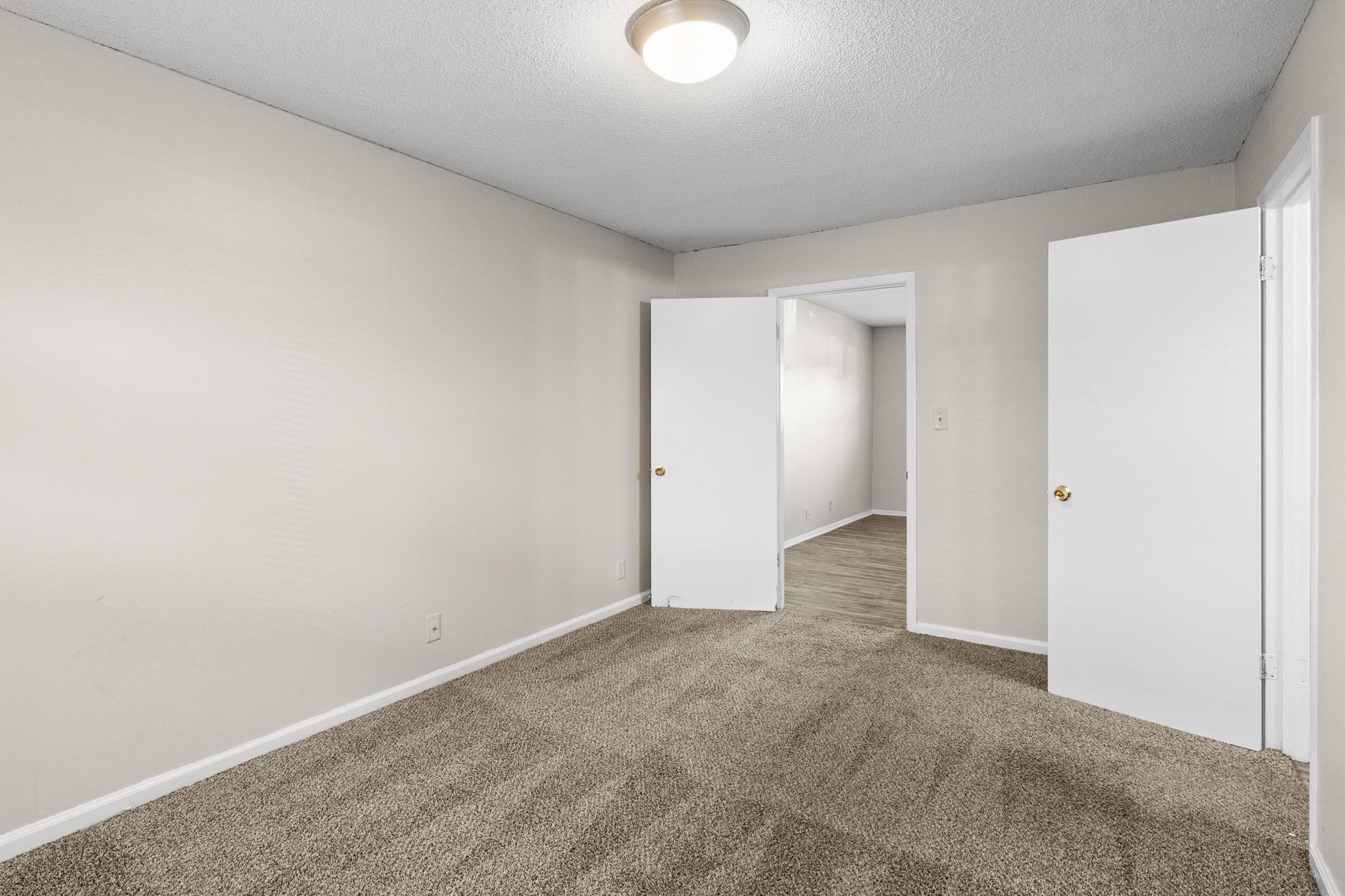 Empty room featuring light beige walls and carpeted flooring. Two doors open to adjacent rooms, and a ceiling light fixture provides illumination. The space is devoid of furniture, emphasizing the clean and simple layout.