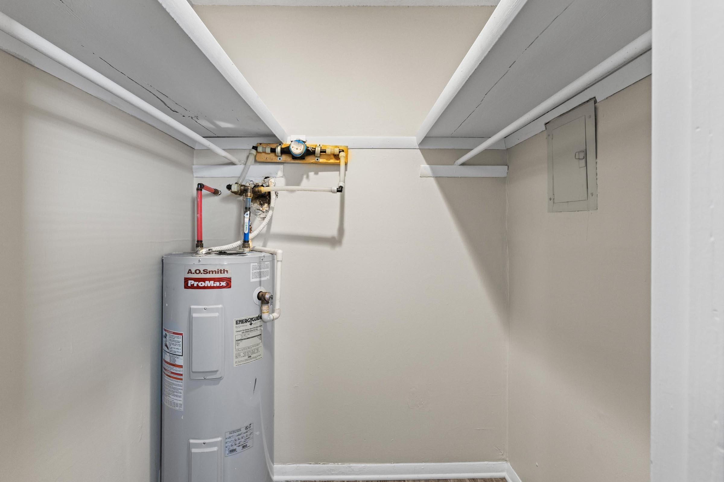 A small, empty storage closet featuring a water heater against one wall. The water heater is silver with a label and pipes attached. The walls are painted a neutral color, and there are white shelves overhead. The floor is bare and the space is well-lit.