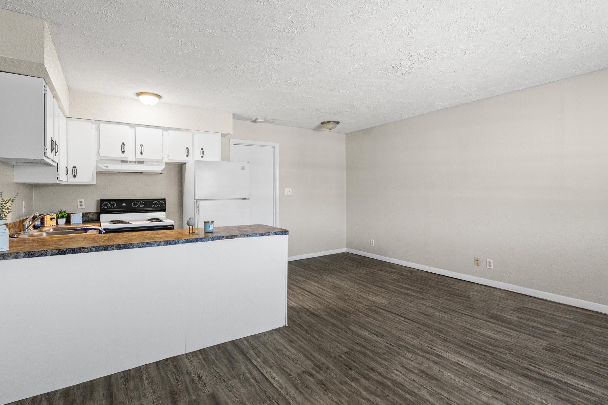 A modern kitchen and living area featuring white cabinets, a countertop with a small sink, and a white refrigerator. The floor is a wood-like laminate, and the walls are painted in a light neutral color. There is a window, allowing natural light into the space. The area is uncluttered and ready for furnishings.