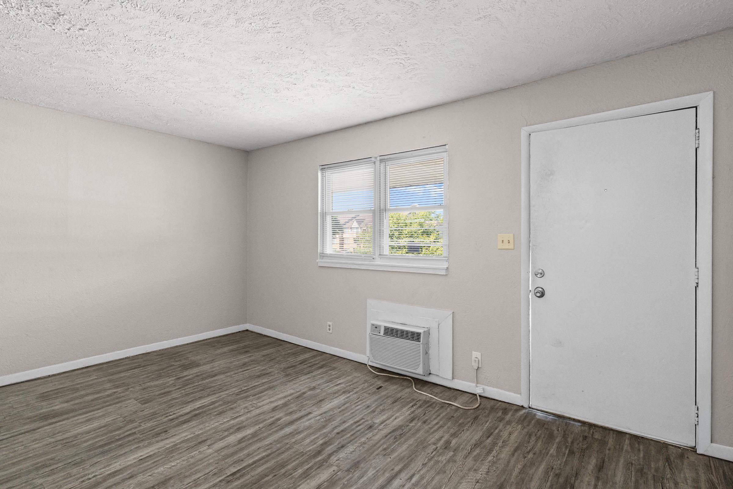 A small, empty room with light beige walls and wood-like flooring. There are two windows allowing natural light, an air conditioning unit on the wall, and a simple white door leading outside. The room appears clean and ready for furniture placement.