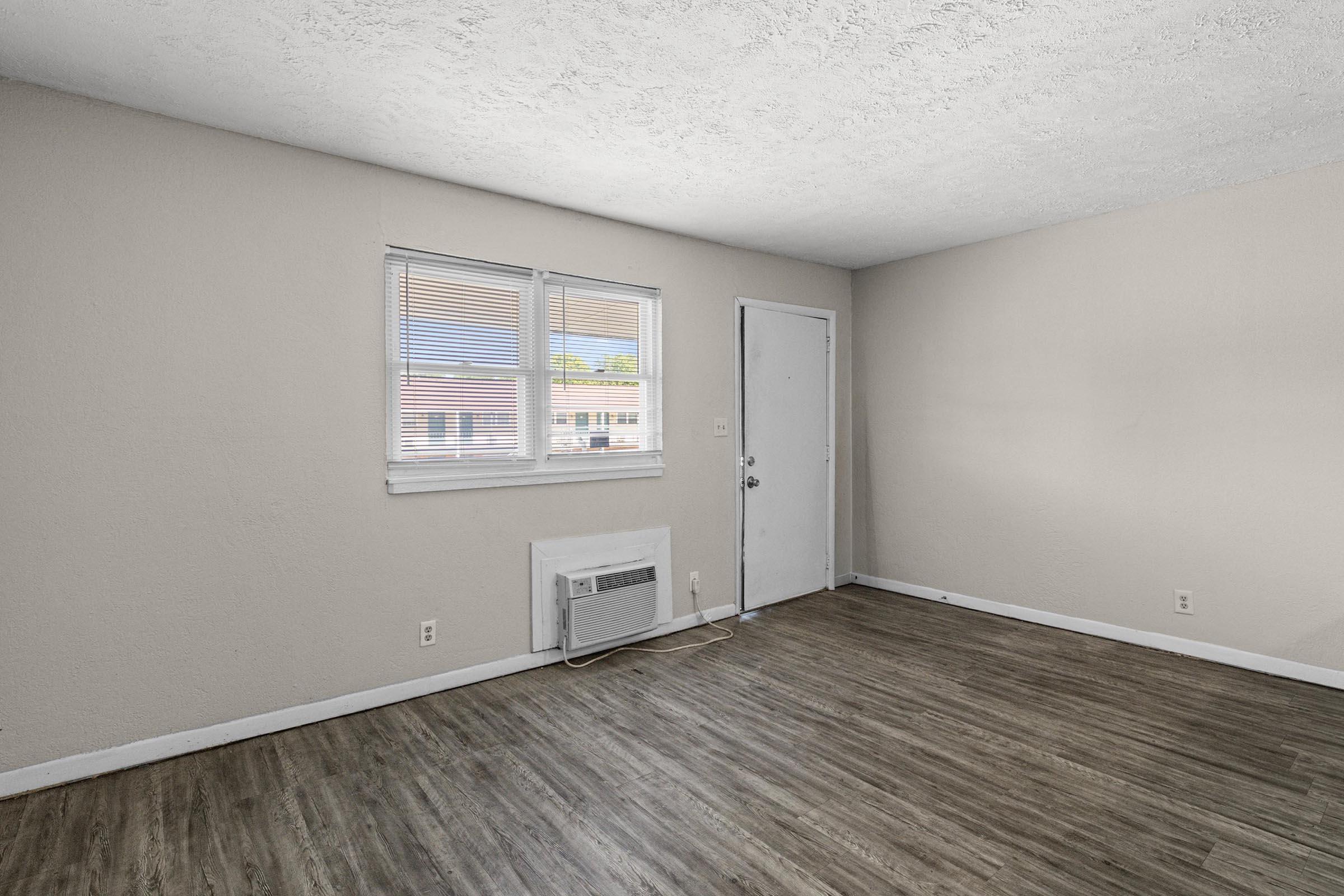 Empty room with light-colored walls and a wooden floor. Two windows allow natural light to enter, and an air conditioning unit is installed on one wall. There's a solid door leading outside, and the overall space appears clean and uncluttered.