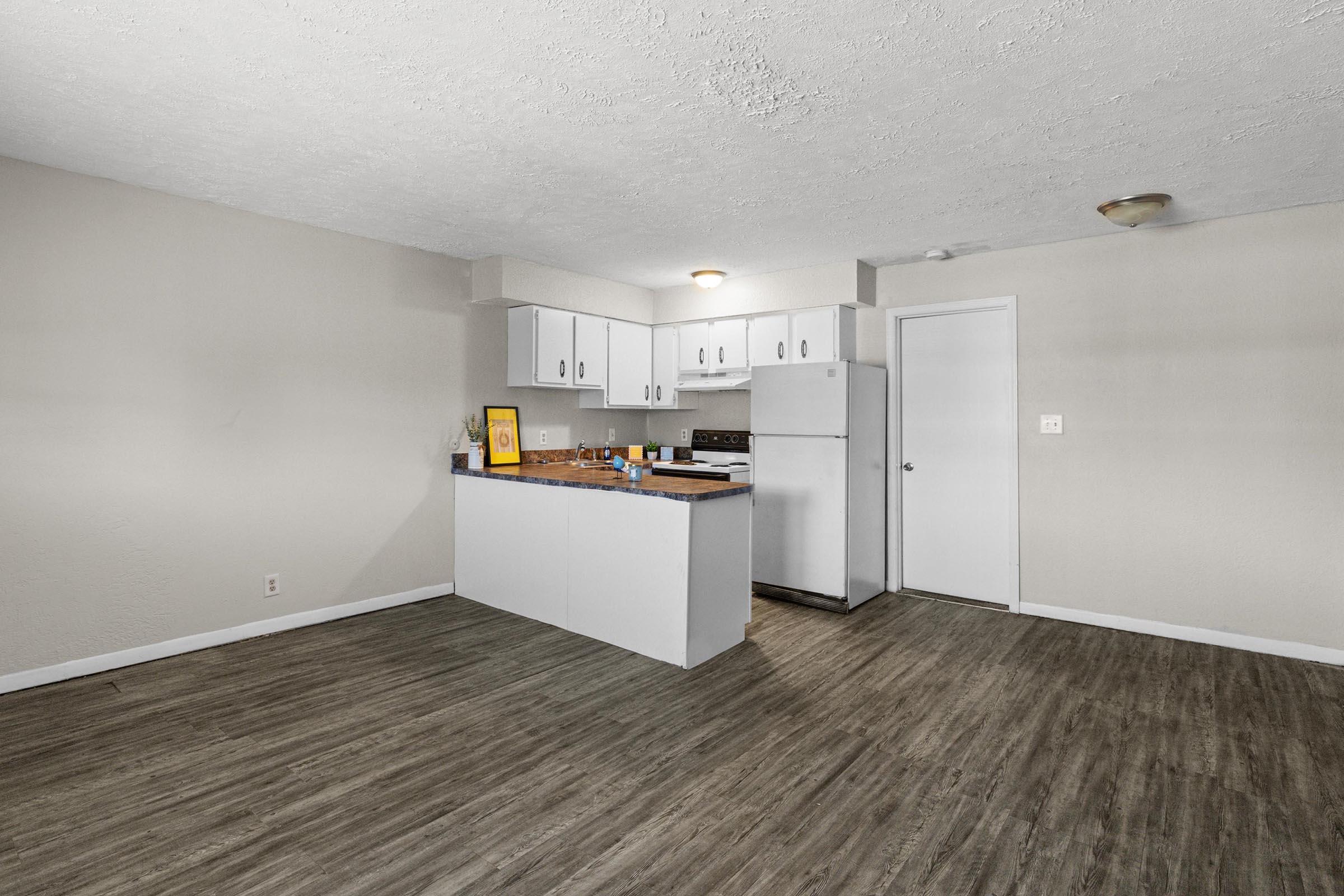 A modern kitchen with a white cabinetry and countertop, featuring a refrigerator, stove, and sink. The open space leads to a well-lit living area with light wood-like flooring. Neutral-colored walls and a simple light fixture enhance the contemporary feel of the room.