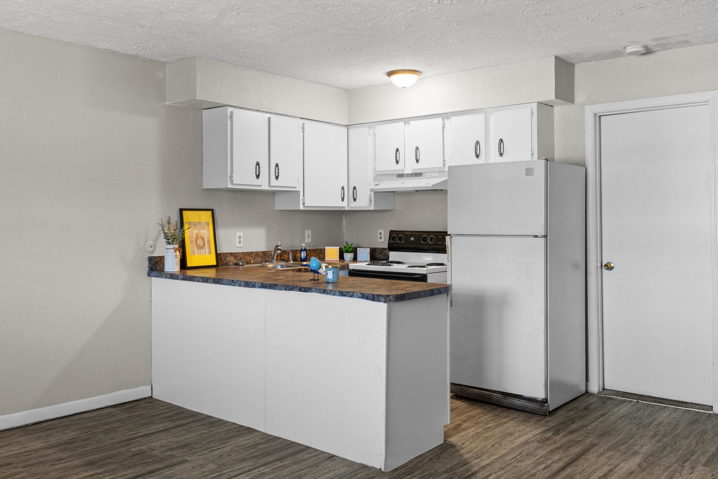 A modern kitchen featuring white cabinets, a marble countertop, and a stainless steel refrigerator. There's a framed picture on the counter, with a blue bowl and some decorative items. The floor is light wood, and the walls are painted a neutral color, creating a bright and airy atmosphere.