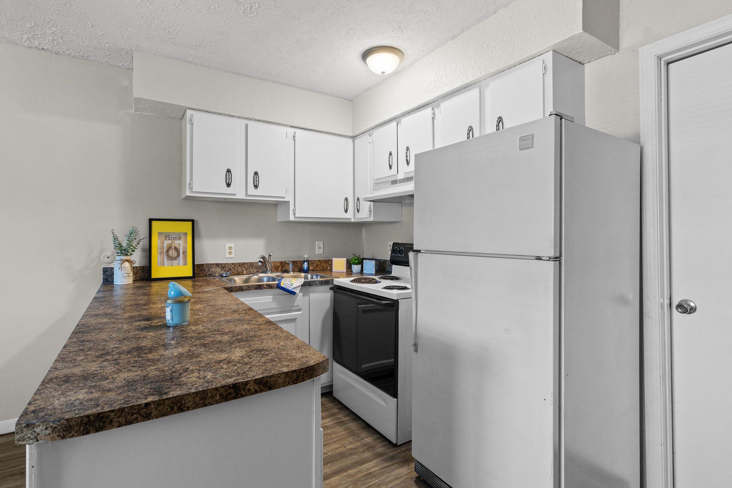 Bright kitchen featuring white cabinets, a beige countertop, and modern appliances. There's a refrigerator, stove, and a small sink area. A blue cup and some kitchen supplies are visible on the countertop, along with a framed picture on the wall. The space has a neutral color scheme, creating a clean and inviting atmosphere.