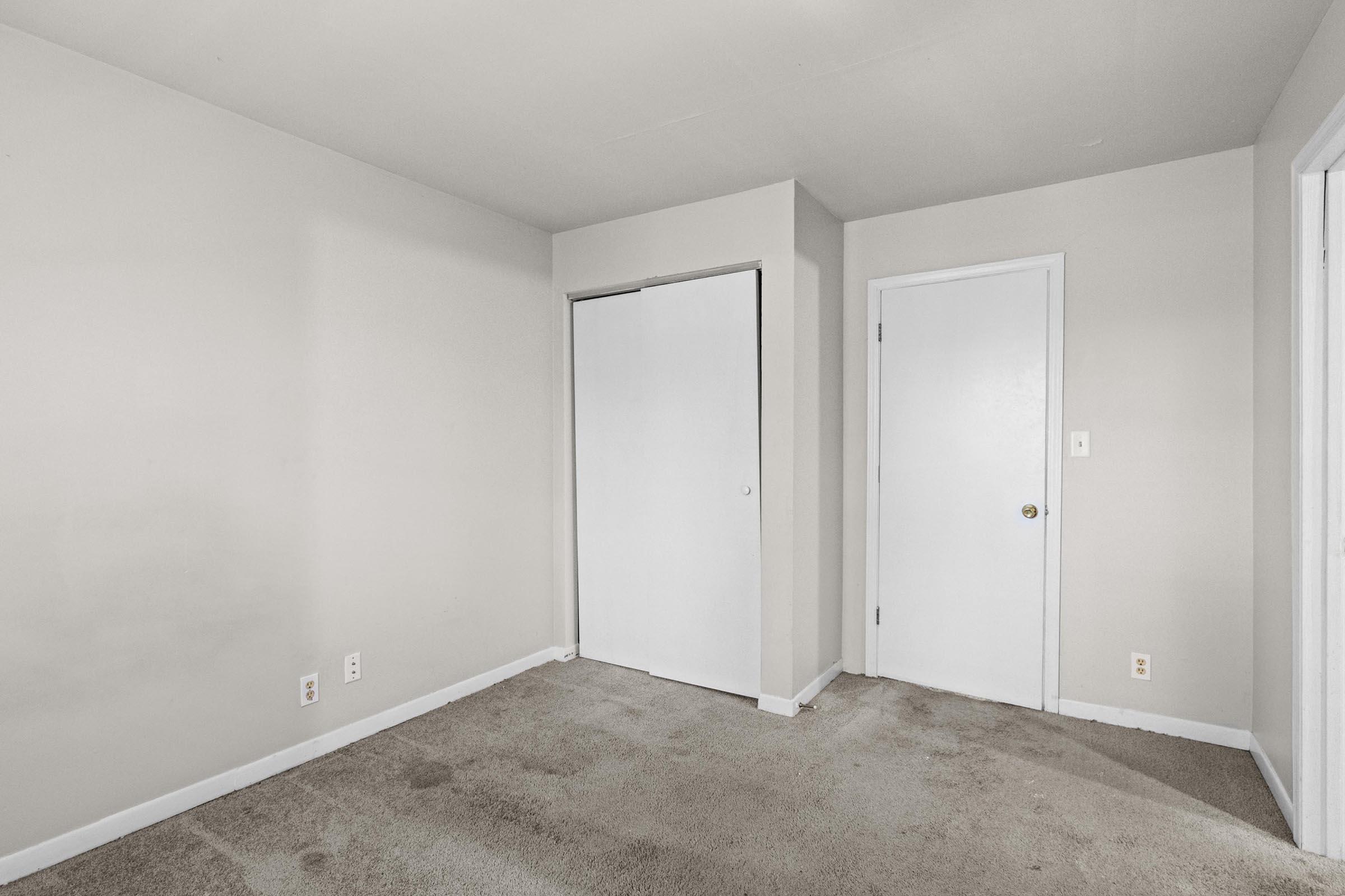 A blank, empty room with light beige walls and a light brown carpet. There are two closed doors on the left and right, both white with simple round knobs. The room is well-lit, highlighting its minimalistic and uncluttered design.