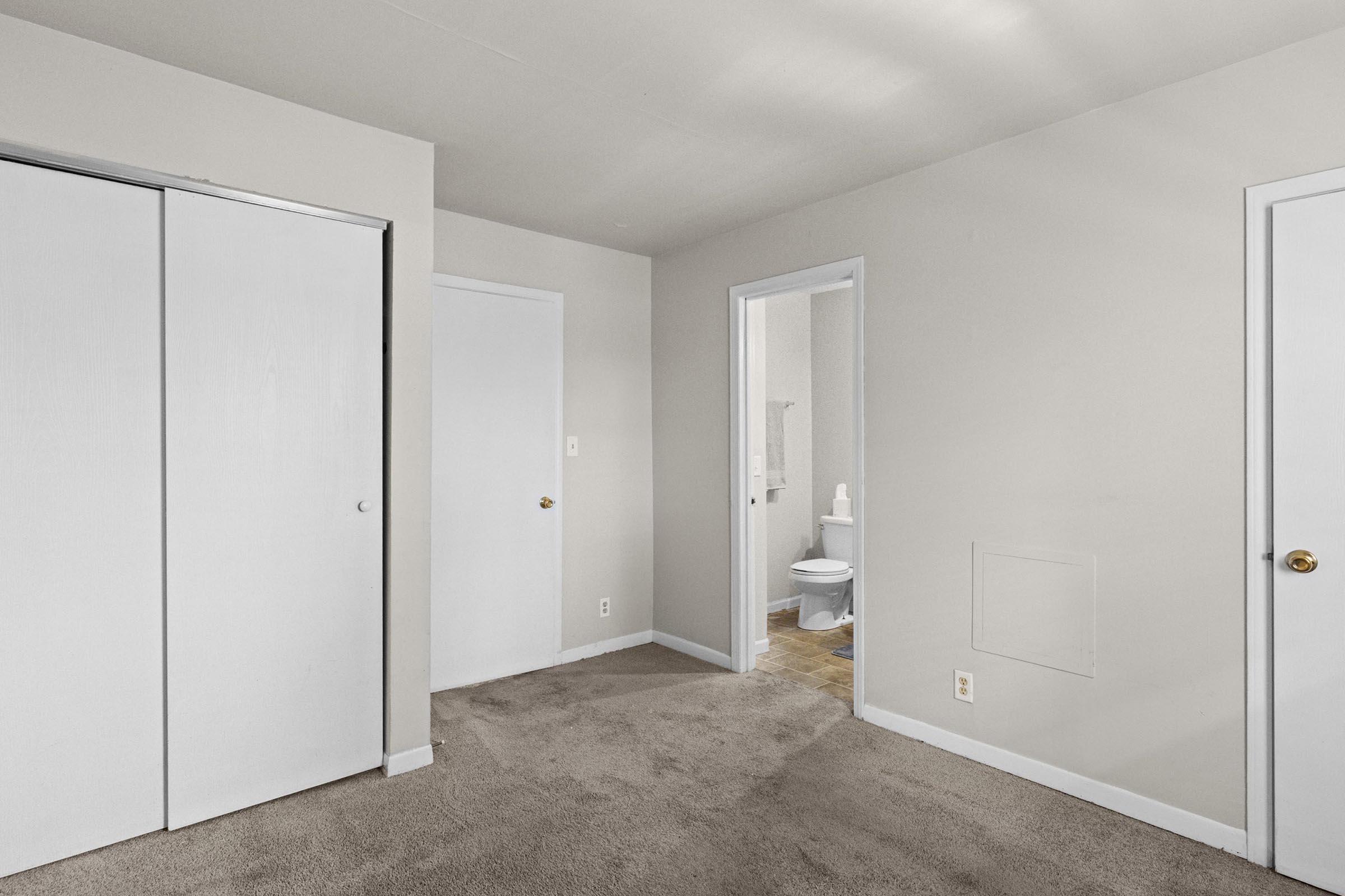 A light-colored room with neutral walls and carpet, featuring two white closet doors. In the background, a doorway leads to a bathroom visible with a toilet. The lighting is soft, and the space appears well-maintained and empty, suitable for furnishing.
