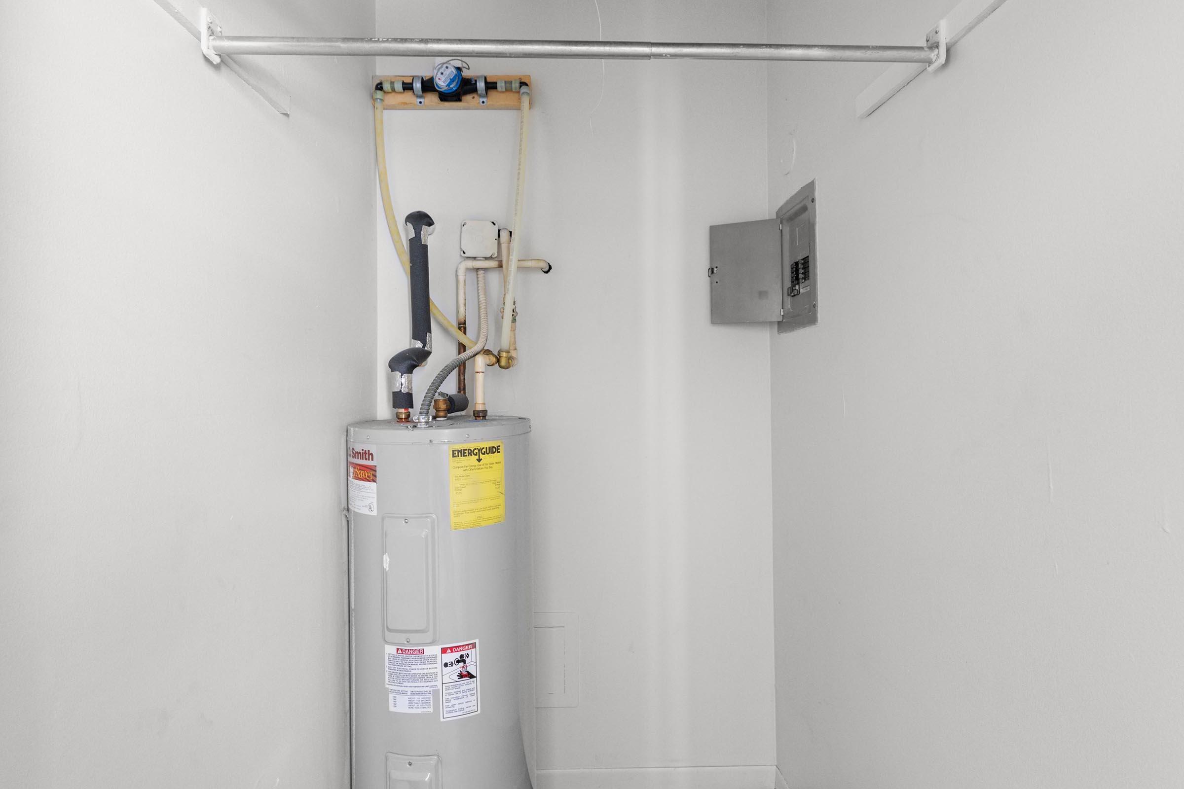 A gray water heater is installed in a small, plain room. It features pipes and a temperature gauge on top. Above the heater, there is a pipe ladder for support. A gray electrical panel is mounted on the wall beside the water heater. The walls are white and the floor is bare.