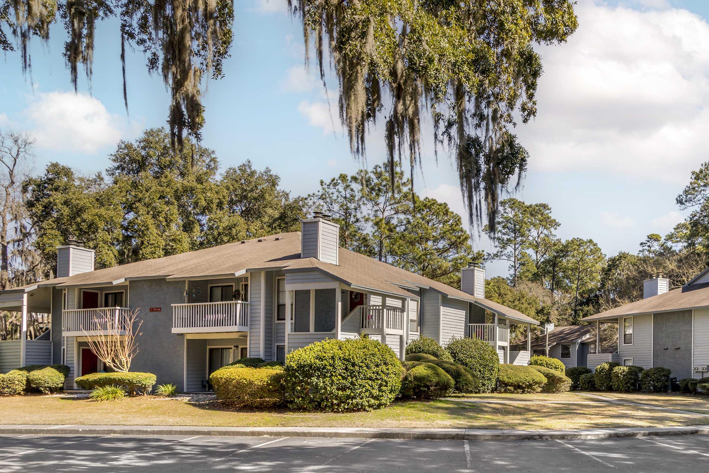 A two-story residential building surrounded by lush greenery and trees, featuring balconies on the upper floor. Spanish moss hangs from the trees, and there are well-maintained shrubs in the foreground, creating a peaceful suburban atmosphere. Bright blue sky and fluffy clouds enhance the scenic view.