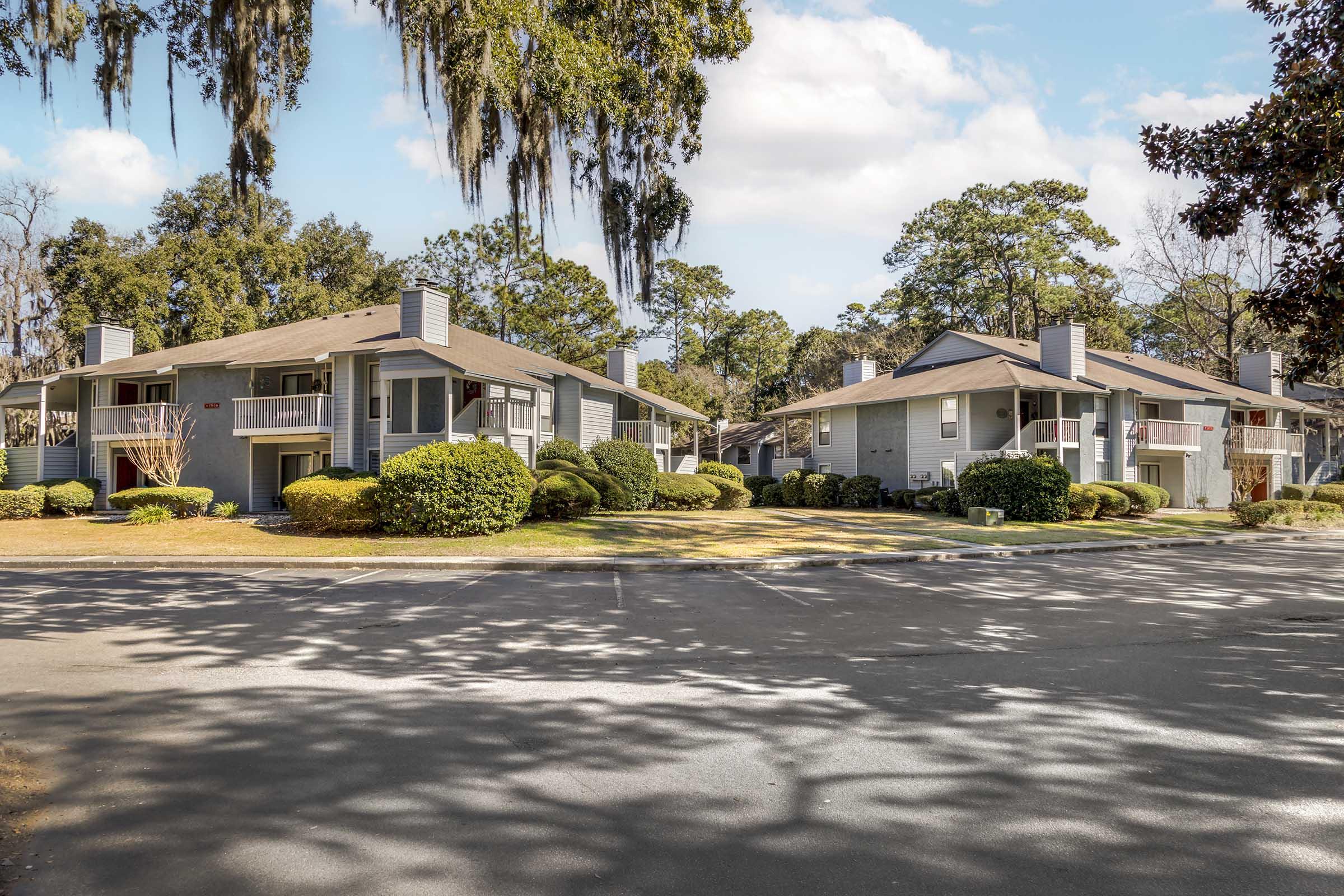 A row of two-story apartment buildings surrounded by lush greenery and neatly trimmed shrubs. The scene features a paved parking area in front of the buildings, with tall trees casting shadows on the ground. The sky is partly cloudy, creating a bright and inviting atmosphere.