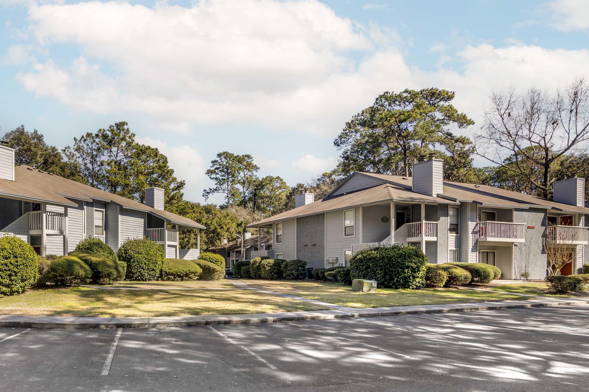 Two-story apartment buildings surrounded by well-manicured lawns and shrubs under a partly cloudy sky. The scene shows a parking area in the foreground, with pathways leading to the entrances of the buildings, creating a tranquil residential atmosphere.