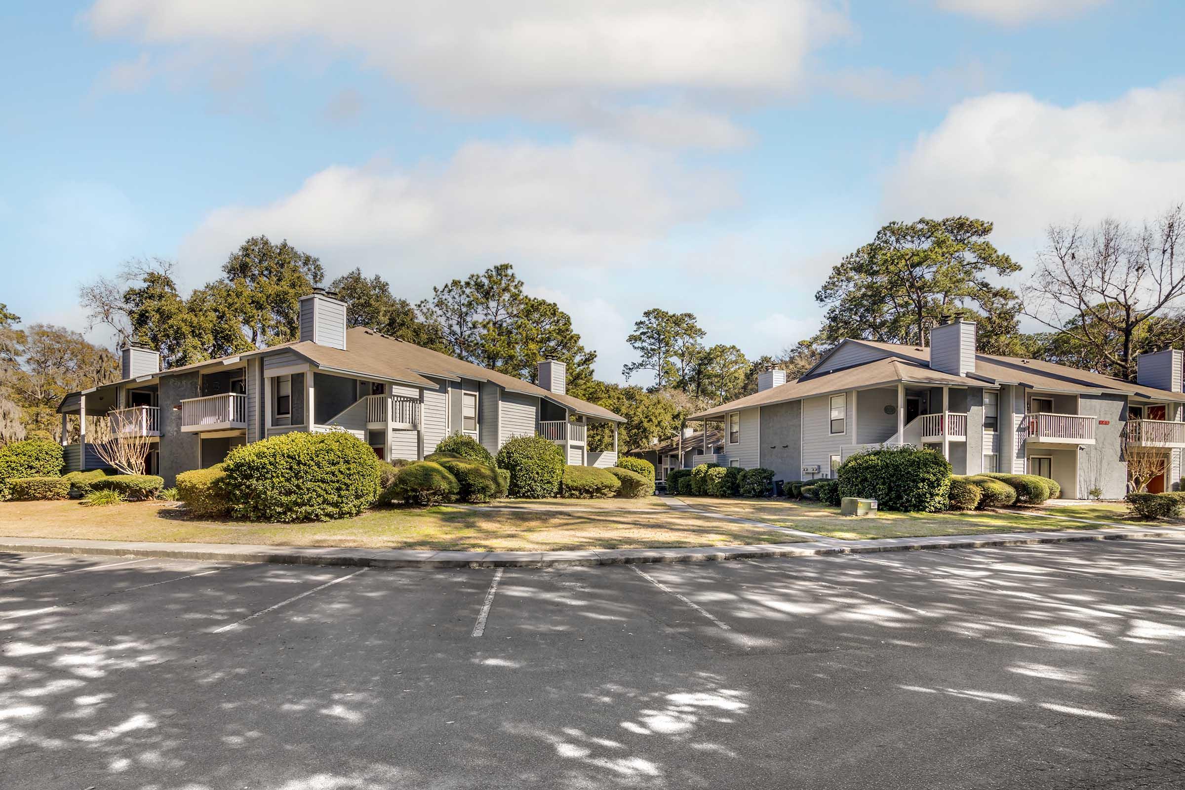 Two apartment buildings surrounded by neatly trimmed bushes and trees, set in a grassy area. The buildings feature sloped roofs and balconies. A paved parking area is visible in the foreground, with a clear blue sky above.