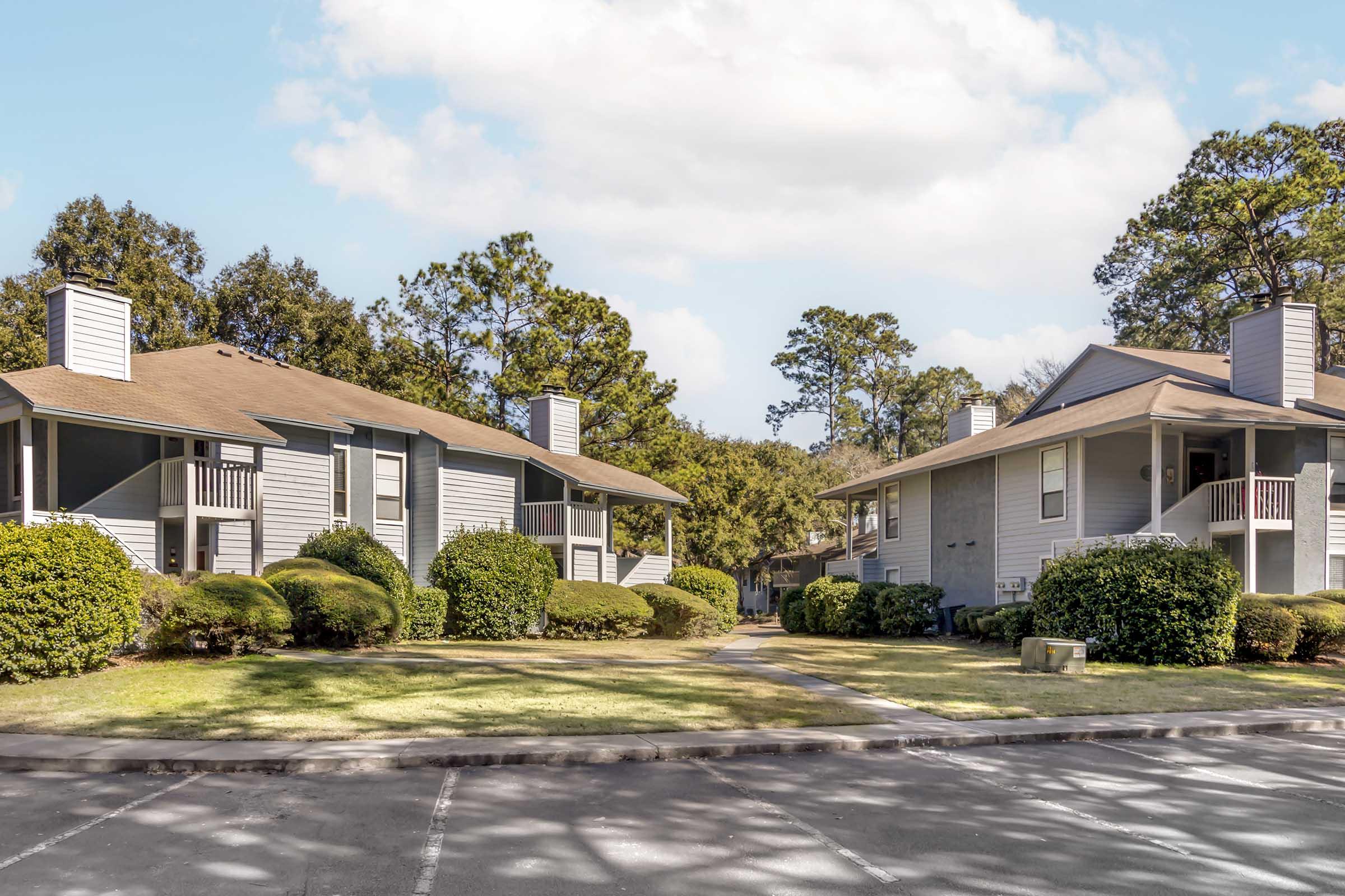 Two residential buildings in a landscaped area with green bushes and trees. The buildings are gray with balconies and sloped roofs, surrounded by a well-maintained lawn. A pathway leads through the area, and the sky is partly cloudy.