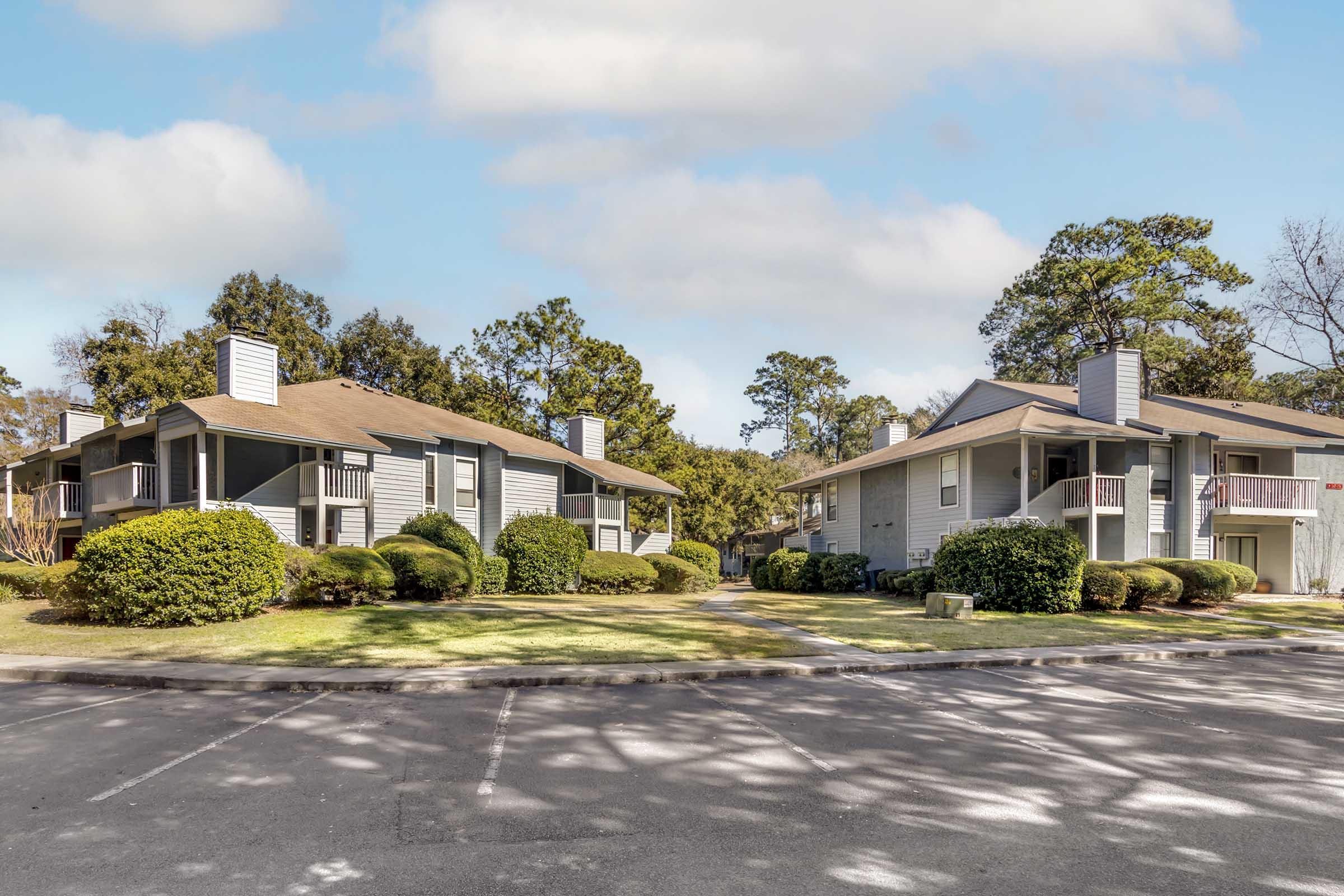 Two residential buildings in a landscaped area with green lawns and shrubs. Each building features a sloped roof, balconies, and a parking area in the foreground. The scene is set under a clear blue sky with a few clouds, and trees are visible in the background.
