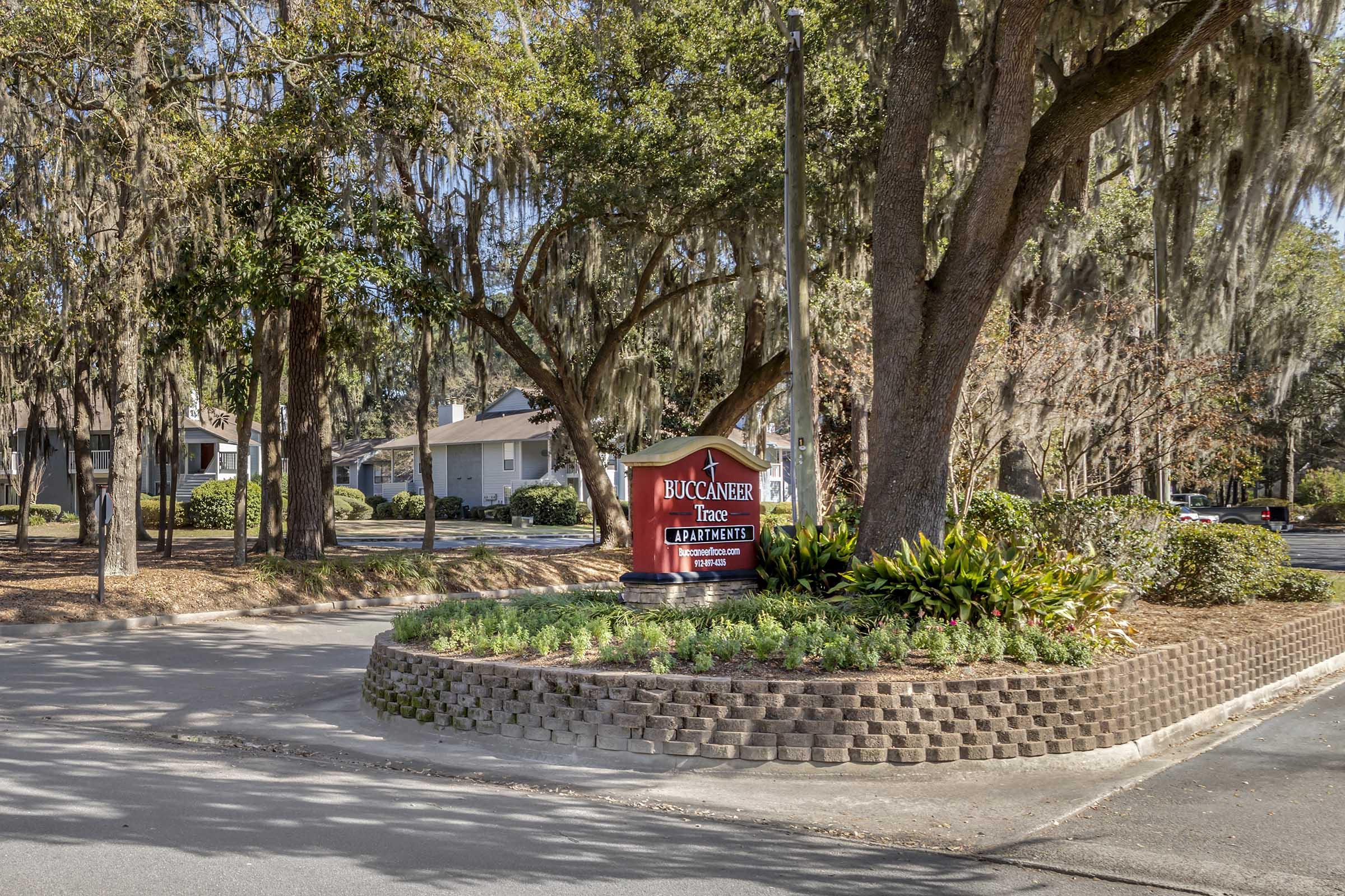 A red entrance sign for Buccaneer Townhomes is visible at the center of a roundabout, surrounded by landscaped greenery and mature trees. The residential area features well-kept homes in the background, with moss hanging from the branches, creating a serene neighborhood atmosphere.