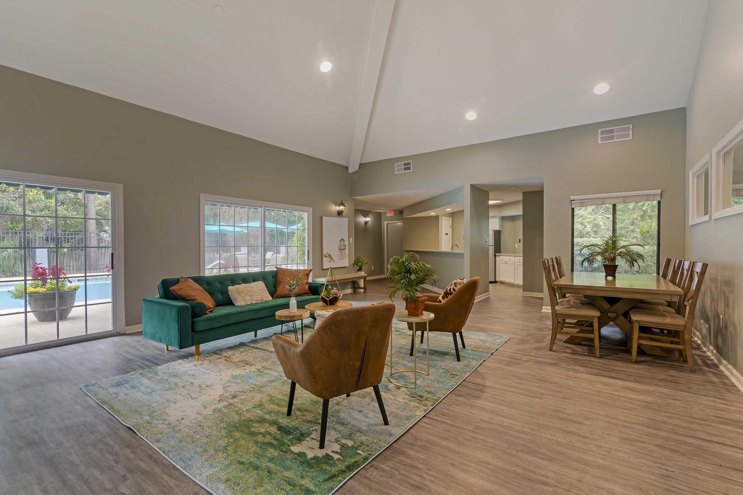 Interior of a modern living space featuring a teal sofa, two brown armchairs, and a wooden dining table. Large windows allow natural light and overlook a pool area. The walls are painted in a soft green, while the floor is a light wood finish. Decorative plants and a rug add warmth to the room.